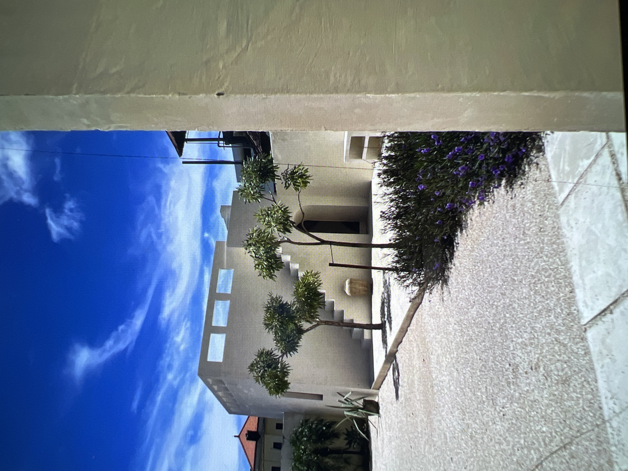 The image depicts a modern courtyard with minimalist architecture. The building is white and boxy, with geometric cutouts along the upper facade. Several small, manicured trees are strategically placed in the courtyard, along with a patch of purple flowering bushes. The ground is covered in light-colored gravel, with some larger square paving stones visible. In the background, another building with a red-tiled roof is partially visible. The sky is a vibrant blue with wispy white clouds. A thin, dark wire or antenna is visible stretching vertically in the upper right portion of the frame. The scene is well-lit by natural sunlight, casting shadows on the ground and building. There are no people visible in the image. This image does not show anything that would suggest it is located in Singapore.