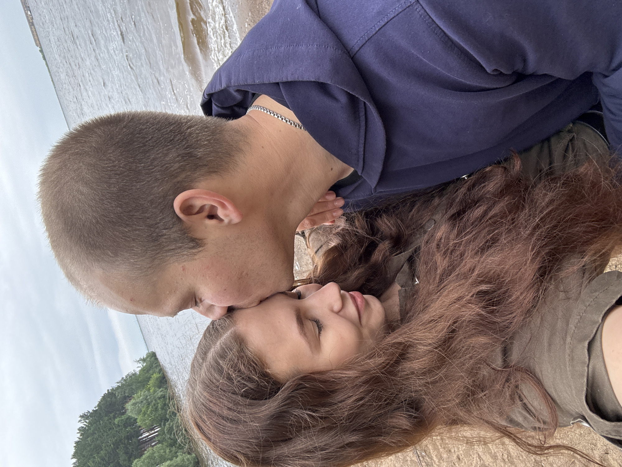 The image captures a close-up of a young couple. A young man, with short brown hair and a silver chain around his neck, is kissing the forehead of a young woman. The woman has long, wavy brown hair and her eyes are closed, with her head tilted back. She is wearing a green jacket. The man is wearing a dark blue hoodie. They are positioned on a sandy surface, with a body of water and a tree line visible in the background. The water appears to be a lake or river, and the trees suggest a natural, outdoor setting. The sky is overcast, indicating a cloudy day. The overall mood of the image is intimate and romantic. No text is visible in the image.