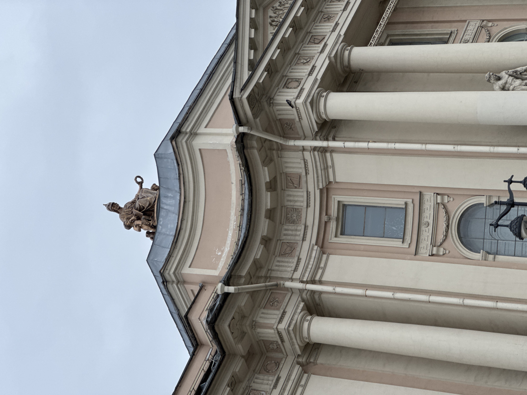 An ornate, multi-story building facade, likely an institutional or palatial structure, is captured from a low-angle perspective against an overcast, light grey sky. The image is rotated 90 degrees counter-clockwise from its natural orientation, showing the building rising vertically on the right side of the frame.
The architecture is characterized by classical elements, featuring large, cream-colored columns, intricate friezes, and cornices. The building's main body is painted a light pink or beige, contrasting with the cream-colored architectural details. Elaborate decorative motifs, including squares, circles, geometric patterns, and laurel wreaths, adorn the entablature sections between levels.
Two sculptures are visible. One, positioned at the apex of the roofline or pediment, depicts a robed figure holding an object, possibly a ring or key. A second, lighter-colored sculpture of a reclining or outstretched human figure is situated on a lower level between two large columns.
Windows of various shapes punctuate the facade: a rectangular window with dark panes and an arched or semicircular window are discernible. An ornate, dark metal lantern or light fixture hangs near the arched window. A metal roof with visible guttering and downspouts runs along the upper edge of the structure. The building shows signs of age and weathering, with some discoloration and wear on its surfaces. There are no people or discernible activities taking place, and no visible text. The overcast sky suggests it is daytime.