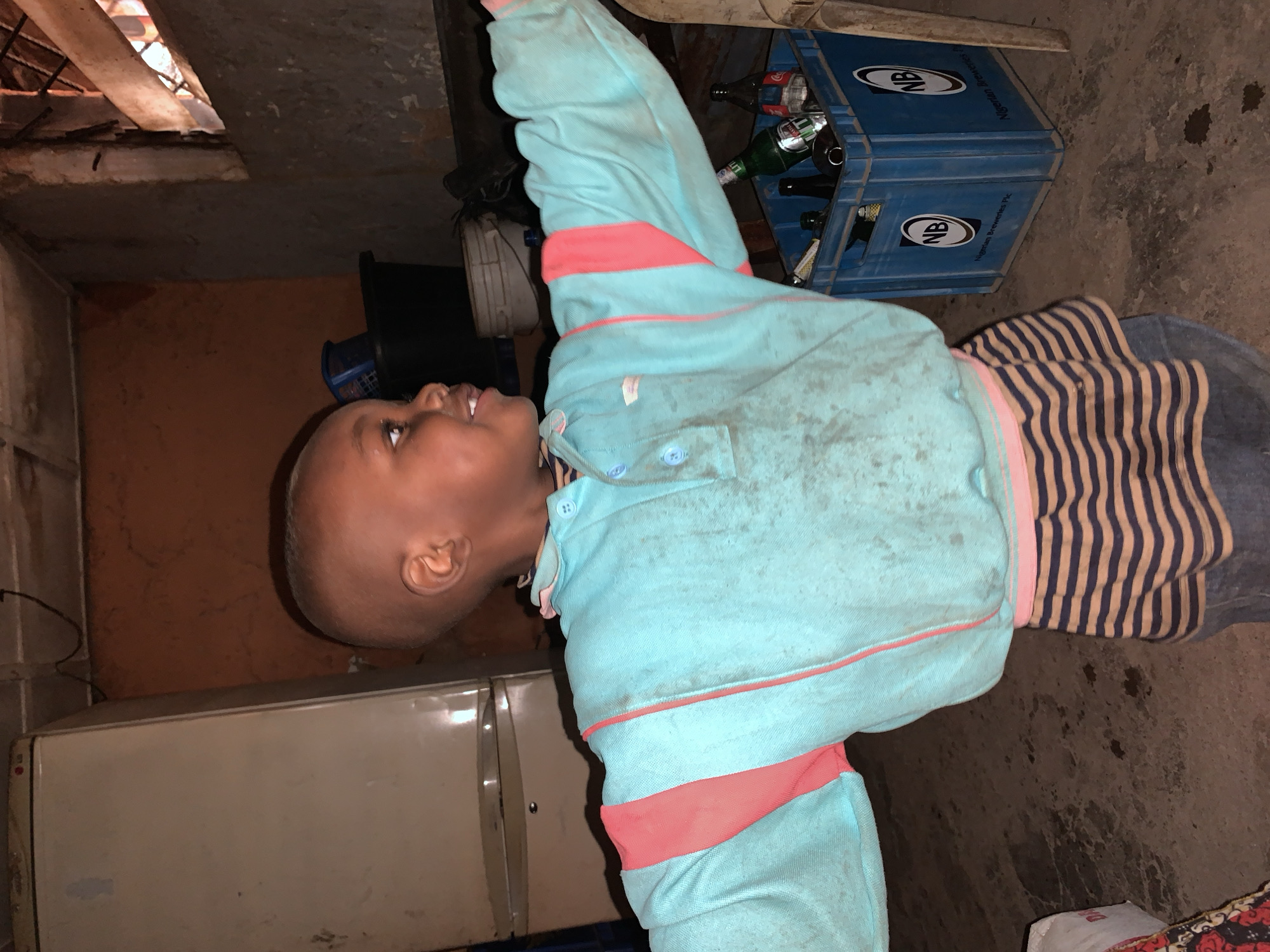 A young child, estimated to be between 4-6 years old, with dark skin and a closely cropped haircut, is captured mid-action within a dimly lit indoor environment in Jos, Nigeria. The child wears a light blue long-sleeved polo shirt, visibly soiled, featuring coral-pink stripes on the sleeves and a matching trim on the collar. Beneath, a garment with dark blue and tan horizontal stripes is visible, paired with dark blue trousers. The child's arms are outstretched horizontally, and the head is turned to the right with a smiling or laughing expression.
In the background to the left, a tall, off-white LG refrigerator is positioned against a reddish-brown textured wall. To the right, a blue plastic crate prominently displaying the "NB" logo and "Nigerian Breweries Plc" text contains multiple empty glass bottles, including a green bottle identifiable as a beer bottle and clear soft drink bottles. Above the crate, a rudimentary window opening with wooden slats and metal bars is visible in a gray concrete section of the wall. The floor is concrete, appearing soiled and uneven. Other background objects include a black bucket with a blue handle and a white bucket. The scene depicts a functional, unadorned interior space.