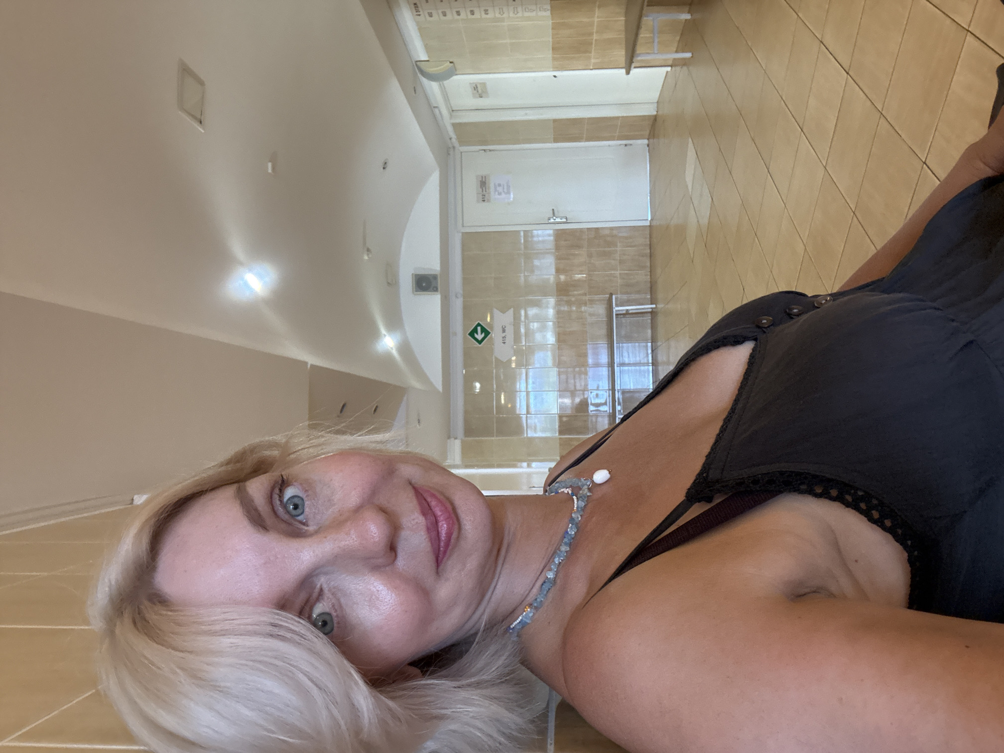 A Caucasian woman with blonde hair and blue eyes is pictured in a selfie. She is wearing a black dress and a beaded necklace. The background appears to be a hallway in a building, possibly a medical facility or waiting area, with tiled walls and floors. There are doors and benches visible. The lighting suggests an indoor setting. The overall impression is a casual, personal photograph. The location is not discernible from the image itself.