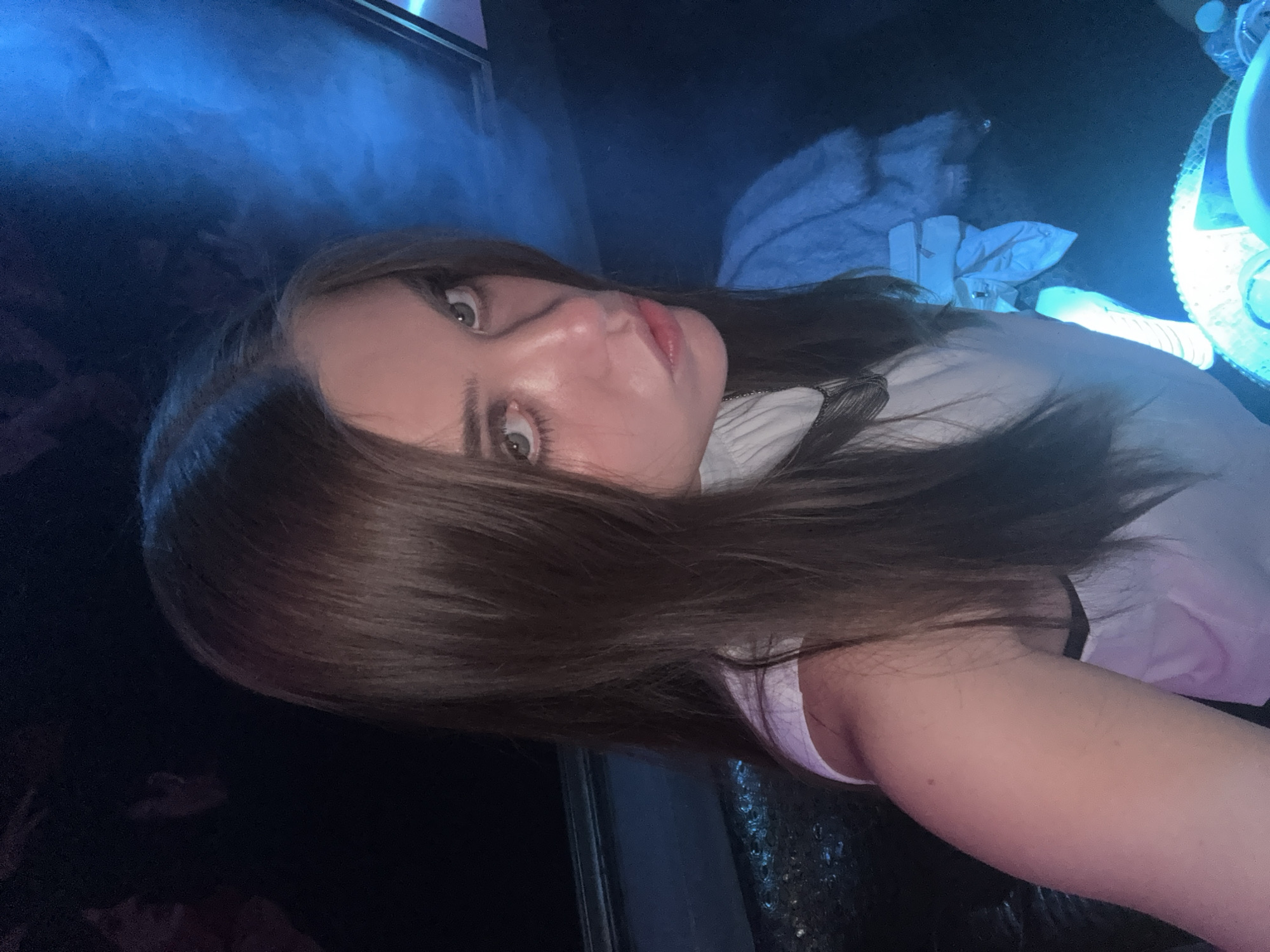 A medium close-up photograph depicts a young female subject with long, straight dark brown hair, parted centrally, and light-colored eyes. She is positioned slightly off-center to the left, looking directly into the camera with a subtle pout. She wears a white, high-neck, sleeveless top; a dark strap is visible beneath her left arm. A multi-strand dark necklace is visible around her neck. Her right arm is bent, partially visible in the lower right quadrant, displaying a small dark mole on the bicep. The environment is dimly lit by strong blue ambient light, creating deep shadows. In the dark background, indistinct figures of at least two other individuals are visible to the left of the primary subject. To the right, a light blue fluffy garment is draped, and a white, folded object is visible below it. A glowing, circular blue light source is partially visible in the lower right foreground, beside a clear plastic water bottle. A faint haze or smoke is present in the upper right background. The scene is consistent with an indoor nightlife setting, such as a club or lounge, in Mexico City, Mexico.