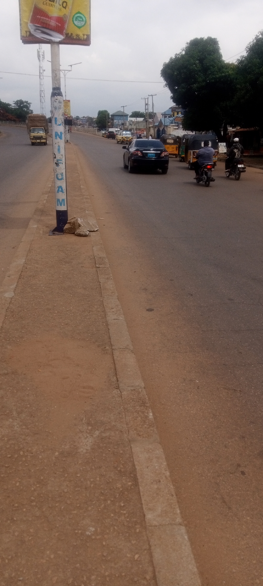 The image captures a street scene in Sabon Tasha, Nigeria. The view is taken from the sidewalk on the side of the road. A light pole with a billboard advertising Guinness is visible. Cars, motorcycles, and tricycles are present on the road. A truck is driving in the far lane. Rocks sit at the base of the light pole.