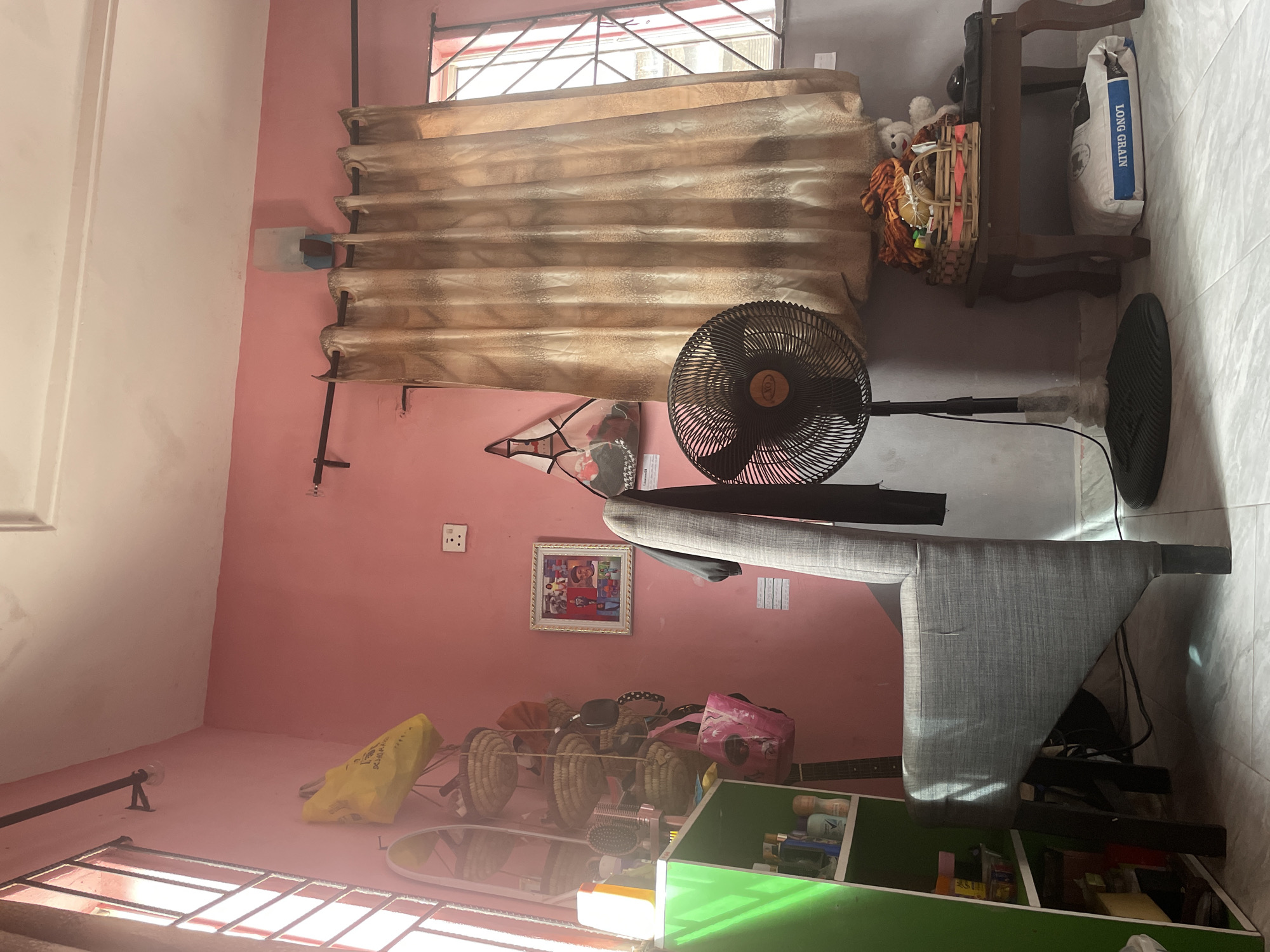 The image depicts an interior scene within a room painted pink, with a view of a window on the left and right sides of the frame. The room is furnished with a grey armchair, a floor fan, a green shelf unit, and a small wooden table.
On the left, a window with white bars lets in bright light, illuminating a yellow bag and several woven baskets hanging on the wall. A green shelf unit is positioned against the wall, filled with various bottles and containers. To the right of the armchair, a floor fan stands near a window covered by beige curtains. On a small wooden table next to the fan, there is a basket filled with items, including what appears to be a stuffed bear and some fabric. Beneath the table, a large white bag with blue lettering is visible.
The overall impression is of a lived-in space, possibly a bedroom or living area. The image does not provide any specific details that would allow for a precise location within Iyafin, Nigeria, beyond it being an interior setting.