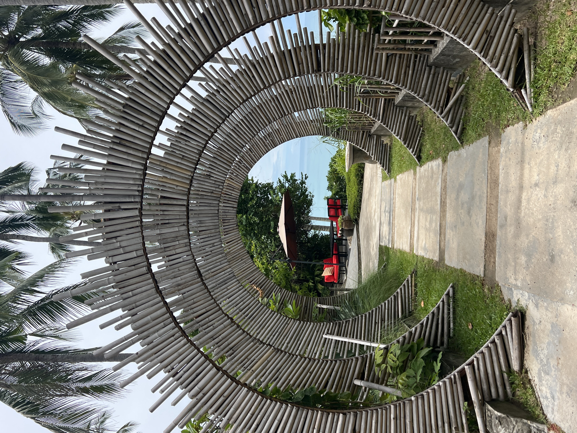 The image captures a pathway framed by a circular bamboo structure, leading towards a seating area. The pathway is made of concrete slabs interspersed with grass. The bamboo structure is a semi-circular archway, constructed from numerous thin bamboo poles arranged radially. Palm trees with their fronds are visible on the left side, indicating a tropical or subtropical location.
Through the bamboo archway, a small seating area is visible. It features two red chairs and a smaller red bench, with a light-colored cushion on the bench. An umbrella, a muted reddish-brown color, is positioned behind the seating. Lush green vegetation surrounds the seating area and the pathway.
The scene suggests a resort or a garden setting, likely in a warm climate given the palm trees and the outdoor seating. The lighting indicates it's daytime, with bright, diffused light suggesting it might be an overcast or slightly hazy day, or the photo was taken during midday. There are no people visible in the image, and no visible text. The overall impression is one of a serene and inviting outdoor space.