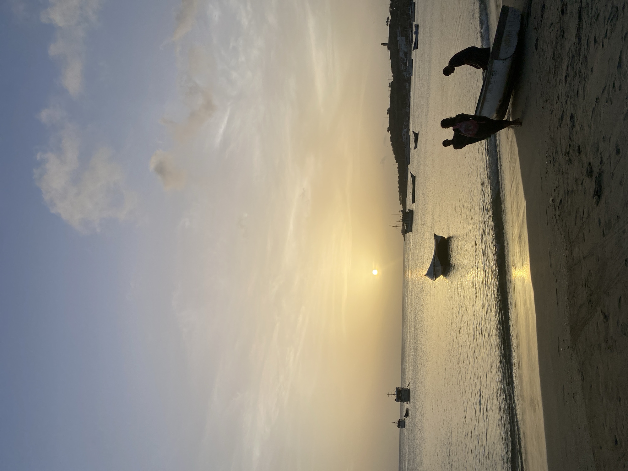 In Juan Griego, Venezuela, at sunset, three silhouetted figures are on a sandy beach near the waterline. Two individuals are pushing a small white boat up the beach, while a third person, wearing a backpack, walks behind them. The sun is low on the horizon, casting a golden glow across the calm water where several other boats are anchored. In the distance, a hilly coastline with scattered buildings and a lighthouse is visible. The sky is a gradient of pale blue and soft yellow, with wispy clouds.