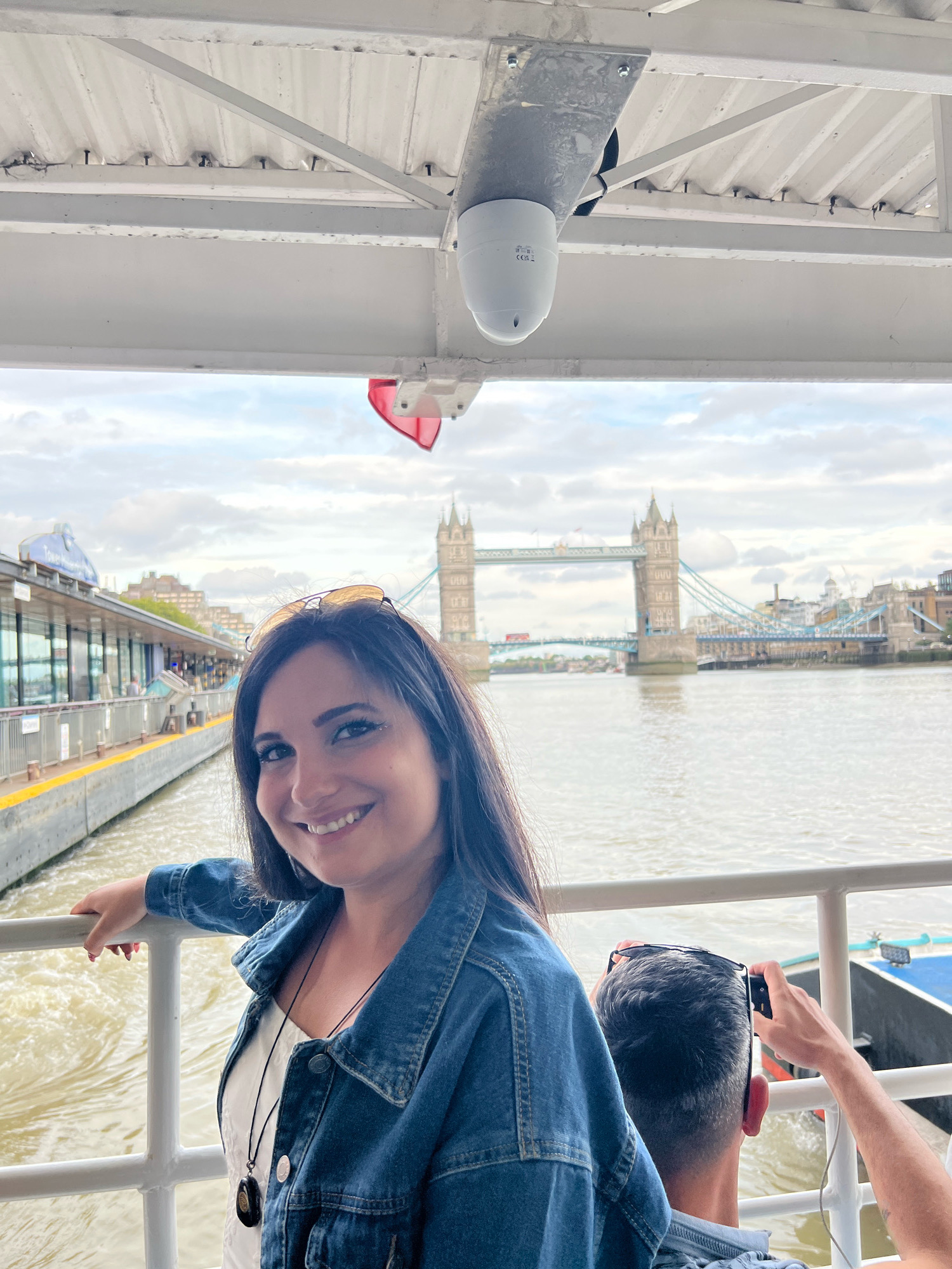 In London, United Kingdom, a woman with dark hair and sunglasses perched on her head smiles at the camera while leaning on a railing on a boat. She wears a denim jacket over a white top with a necklace. Behind her, Tower Bridge is visible across the River Thames. The boat's overhead structure, a security camera, and a red flag are also in the frame. Another person, with a camera in hand, is partially visible in the foreground, taking a photo. The scene is set against a cloudy sky and the backdrop of the London cityscape.