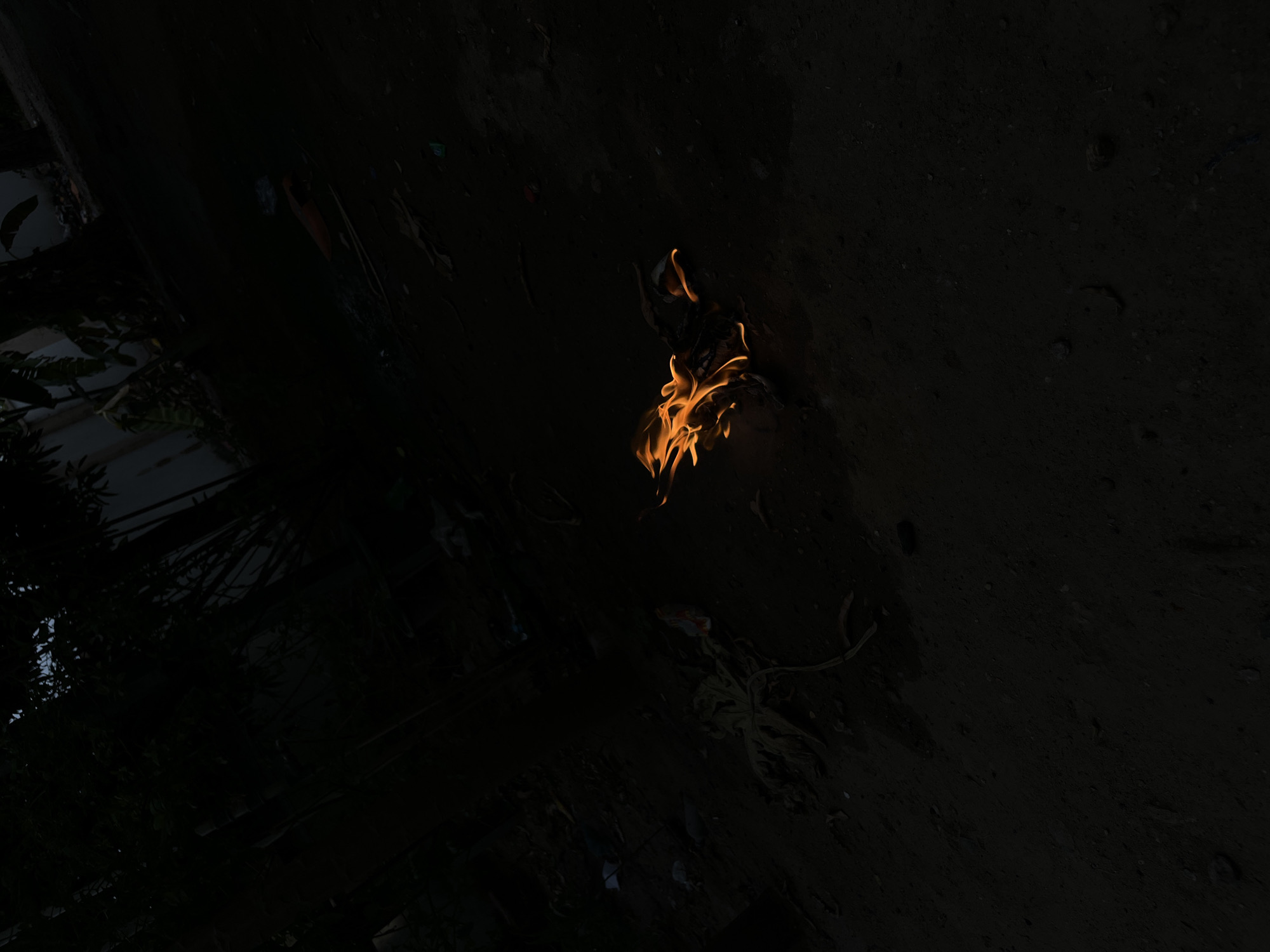 A small fire burns in a dark area. The location is unknown, but the prompt suggests it is in Lokoja, Nigeria. The fire is small and appears to be burning low to the ground. There are no people visible in the image. Other than the fire, the environment is very dark and indistinct. There is a surface, possibly ground or pavement, in the background.
