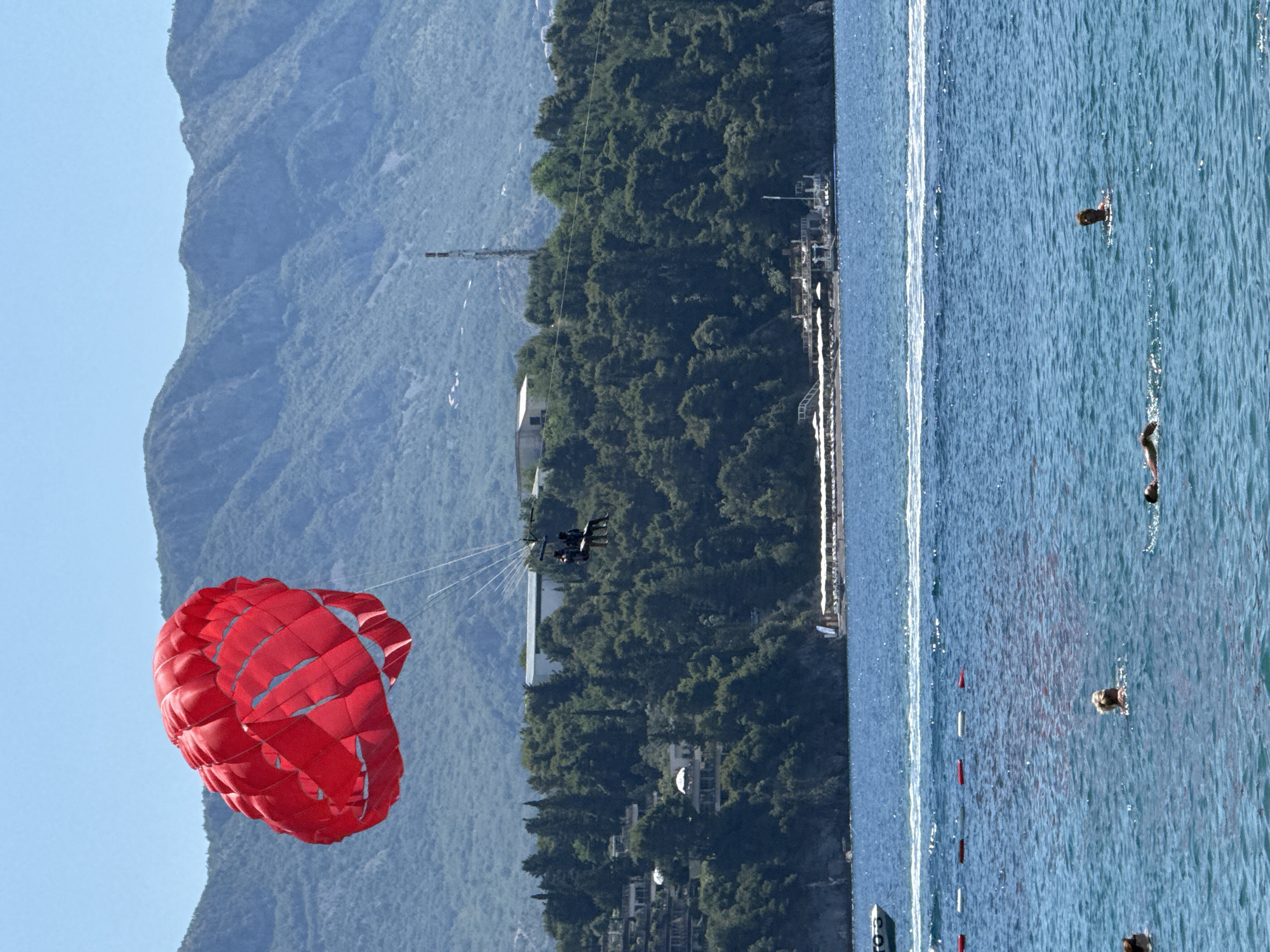 This image depicts a parasailing activity over the Adriatic Sea, near Budva, Montenegro. A bright red parasail is visible in the upper left quadrant of the frame, high above the water. Attached to the parasail are two people seated in a harness. They are positioned over a densely forested hillside dotted with white buildings, characteristic of the coastal architecture found in the region.
The sea is a deep blue, with gentle waves. Several people are swimming or wading in the water closer to the shore. A rope barrier with red and white buoys separates the swimming area from the open water. In the background, a distant mountainous landscape under a clear blue sky suggests a sunny day. A boat is partially visible on the far left, and a structure with white umbrellas, likely a beach club or restaurant, is situated on the shoreline in the middle ground. A tall transmission tower is also visible on the hillside behind the trees. The overall scene conveys a leisurely vacation atmosphere.