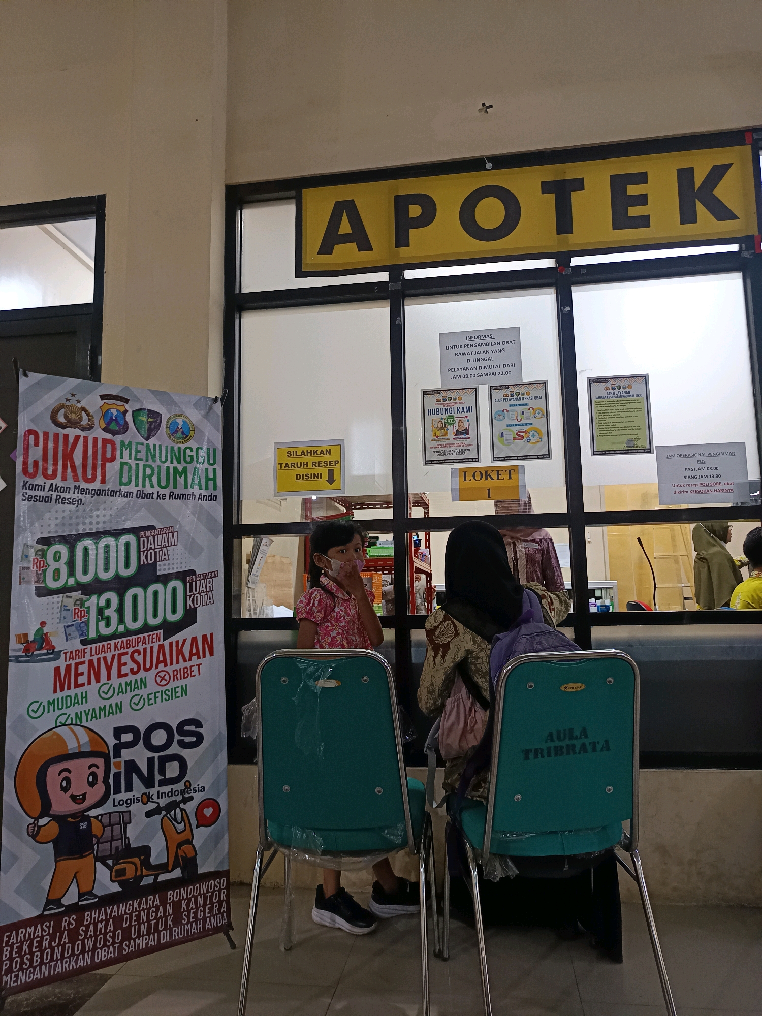 The image shows the interior of an Indonesian pharmacy. A large yellow sign above a counter reads "APOTEK," which means pharmacy in Indonesian. Below the sign, a glass window separates the waiting area from the pharmacy counter. In the waiting area, two green chairs are visible, one of which has the text "AULA TRIBATA" printed on its back. A young girl wearing a mask is seated in the foreground, while two women are standing at the counter. A large banner on the left side of the image advertises a home delivery service for medicine, listing prices for delivery within and outside the city. The banner also mentions "POS IND" and "Logistik Indonesia," indicating the involvement of the Indonesian postal service in the delivery. The overall scene depicts a typical pharmacy setting where customers wait to be served. The banner mentioning "Bondowoso" suggests that this pharmacy is located in the city of Bondowoso, Indonesia.