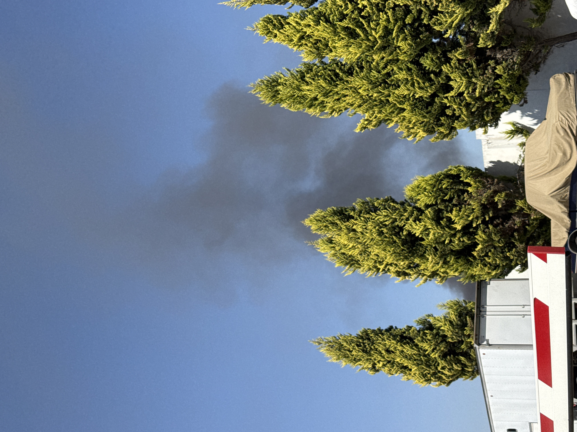 The image captures an outdoor scene under a clear, bright blue sky, with dark smoke visible against the azure backdrop. Three dense, bright green trees, possibly cypresses or junipers, are prominent, their foliage illuminated by daylight. A thick plume of dark grey and black smoke rises and spreads horizontally across the sky, appearing to emanate from a source situated behind the trees or obscured structures.
To the right of the image, a light-colored building or structure, possibly white or light grey, is partially visible behind the foliage. Further right, a large object covered with a tan or khaki-colored tarp suggests equipment or another vehicle. In the bottom right foreground, a section of a white or metallic silver vehicle or trailer is visible, featuring distinctive red and white diagonal safety stripes on what appears to be a rear panel or barrier.
The presence of smoke indicates an ongoing fire or combustion event. The bright, clear sky suggests it is daytime. No people are visible, and there is no discernible text. The scene is set in San Francisco Ocotlán (Ocotlán), Mexico.