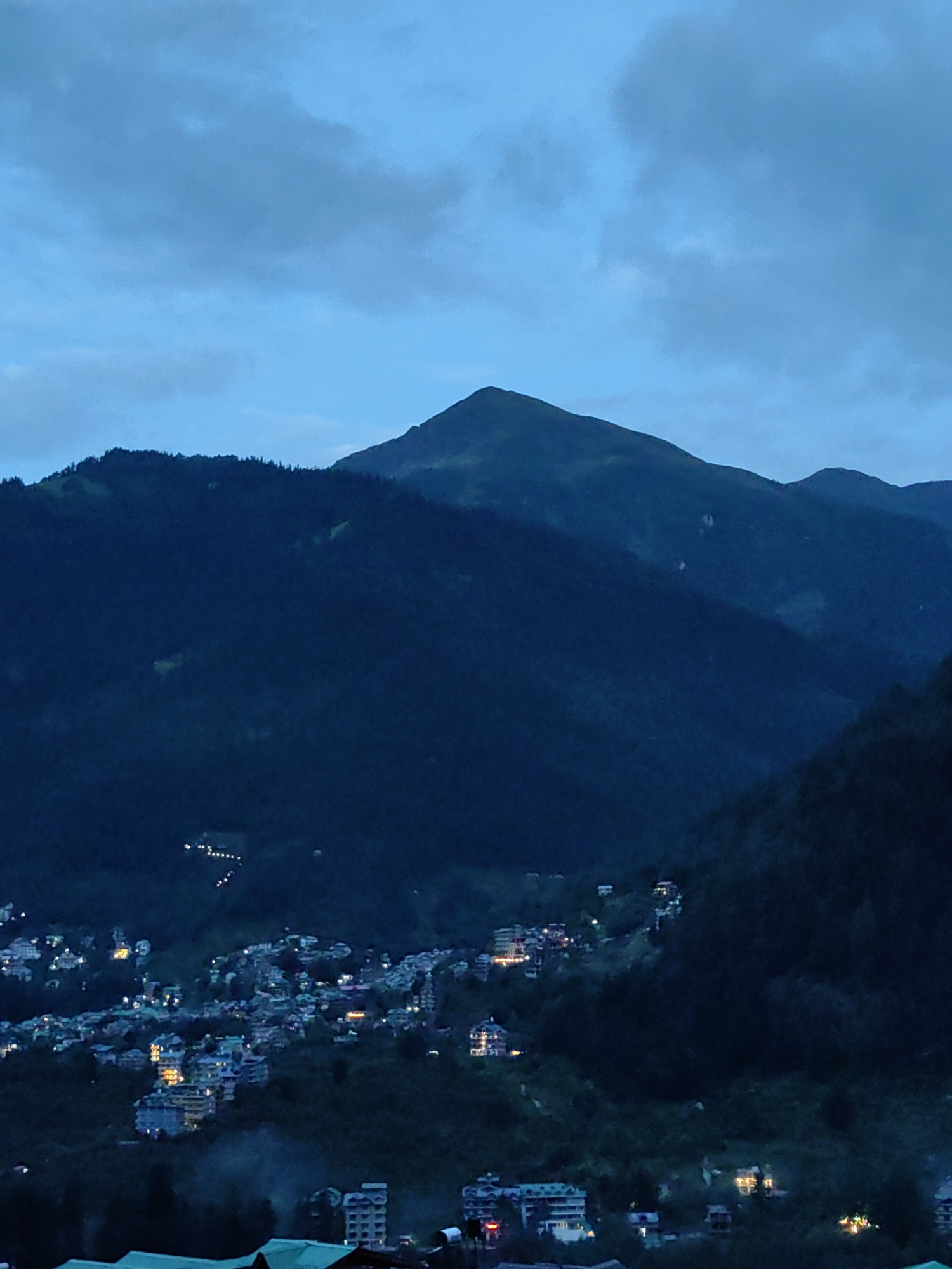 The image captures a dusk scene in Vashisht, India, showcasing a mountainous landscape with a town nestled on the slopes. A prominent mountain peak dominates the background, silhouetted against a twilight sky filled with scattered clouds. The mid-ground reveals a densely populated town, its buildings illuminated by artificial lights, creating a vibrant contrast with the dark mountainside. The buildings appear to be a mix of residential and commercial structures, some featuring multiple stories and balconies. The foreground shows a less dense area with fewer lights, suggesting the edge of the town or undeveloped land. The overall impression is one of a charming mountain village coming alive as night falls, with its lights twinkling against the vastness of the natural surroundings. The area appears to be a popular tourist destination, with numerous hotels and guesthouses visible, indicative of Vashisht's reputation for its hot springs and spiritual ambiance.