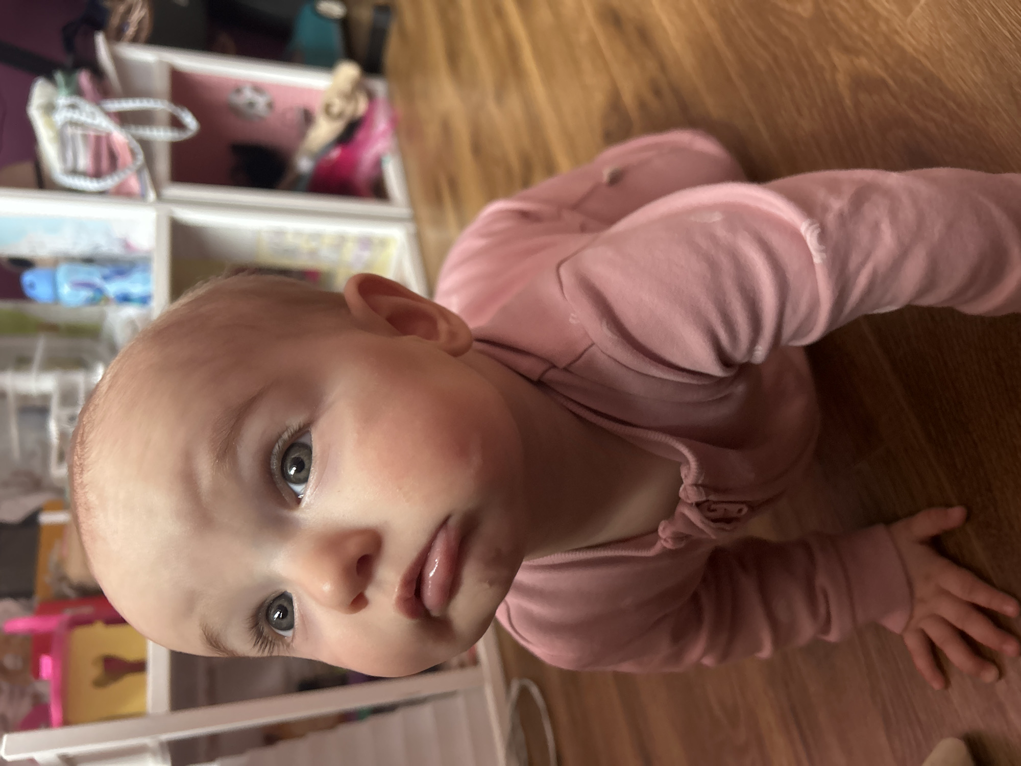 A baby in a pink outfit lies on a wooden floor. The baby's head is close to a white shelf. The location is not discernible as being in Moscow, Russia.