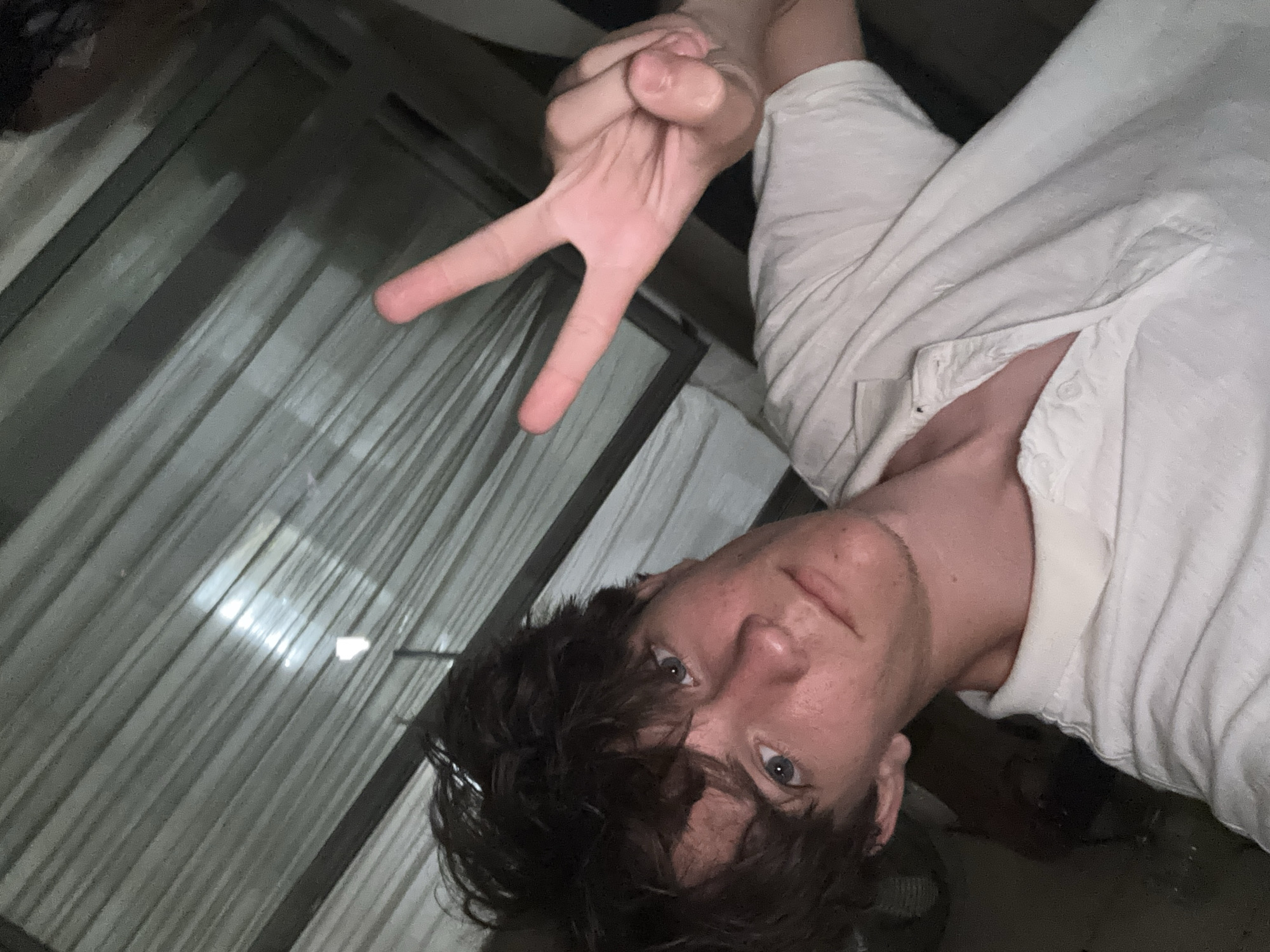 In this close-up, indoor, selfie-style photograph, a young Caucasian male with dark, tousled hair and light blue eyes is pictured. He is wearing a white, collared polo shirt and is holding up his right hand in a peace sign. The background consists of vertical blinds, which are illuminated by a light source, creating diagonal lines of light and shadow. The overall lighting is dim, suggesting an evening or nighttime setting. There is no identifiable information within the image that suggests a specific location in the city of Cupra Marittima, Italy.