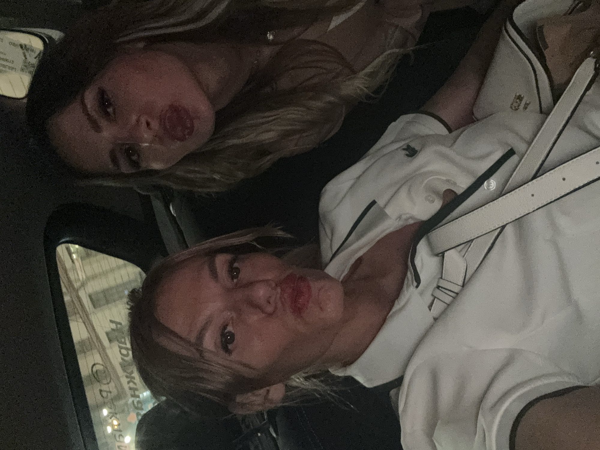 The image depicts two women posing for a selfie inside a vehicle at night. The woman in the foreground, positioned on the left, has blonde hair with bangs and is wearing a white collared shirt with dark trim, featuring a green crocodile logo (Lacoste) on the left chest area. A white shoulder bag strap crosses her body. She has red lipstick and her lips are puckered. The second woman, positioned to the right and slightly behind, also has blonde hair, red lipstick, and puckered lips, looking towards the camera. She wears a light-colored top and a delicate silver necklace. The car's dark interior is visible, including headrest and upholstery. In the reflection on the vehicle's rear window, a horizontally mirrored street sign clearly displays the Cyrillic text "НАБ. КРЮКОВА" (Nab. Kryukova), which translates to "Kryukov Embankment," specifically identifying the location as the city of Saint Petersburg, Russia. Other blurry urban lights and building outlines are also visible in the reflection.
