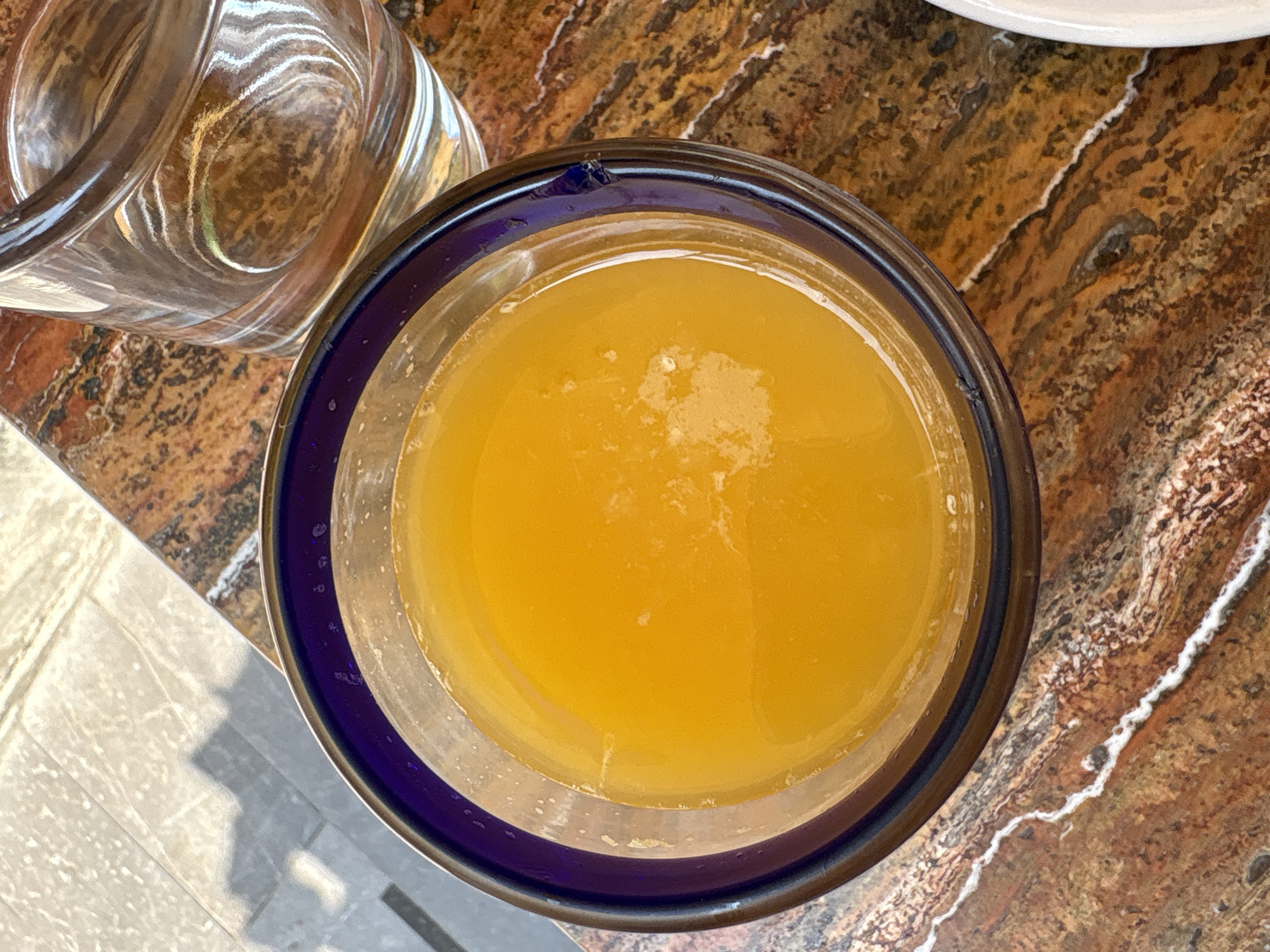 A top-down view depicts a glass of orange juice on a table with a brown and beige pattern, resembling marble or granite. A clear glass with water sits beside the orange juice. Part of a white plate is visible at the top right. The table surface suggests an indoor setting, possibly a restaurant or cafe. The lack of distinct architectural elements or identifying landmarks prevents definitive determination of the exact location within Mexico City, Mexico, but the presence of a glass of orange juice implies it could be a dining establishment catering to tourists. No people are in the frame.