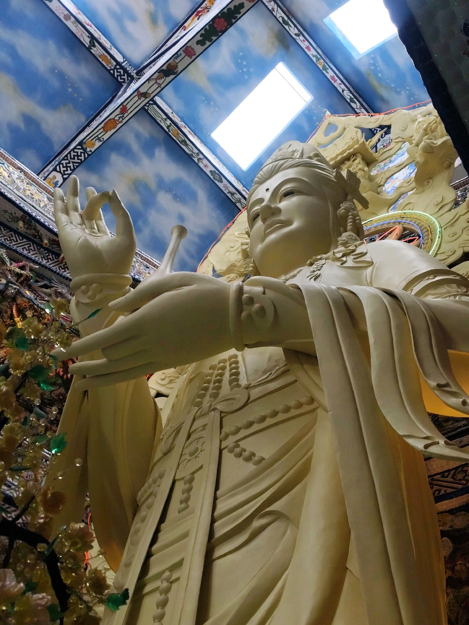 This is a low-angle, full-shot photograph of a large, cream-colored statue of a deity, possibly Guan Yin, in the city of Xuan Thanh, Vietnam. The statue is made of a smooth, matte material and is intricately detailed with flowing robes, ornate jewelry, and delicate hand gestures. The deity's face is serene, with eyes closed and a gentle smile. The statue is positioned in front of a richly decorated ceiling with a celestial motif of blue clouds and stars, and geometric patterns. Two rectangular skylights are visible in the ceiling, illuminating the scene from above. To the left of the statue, a decorative tree with yellow and green foliage and small lights is visible, adding a touch of color and texture to the composition. The overall impression is one of grandeur and spirituality, evoking a sense of peace and reverence.