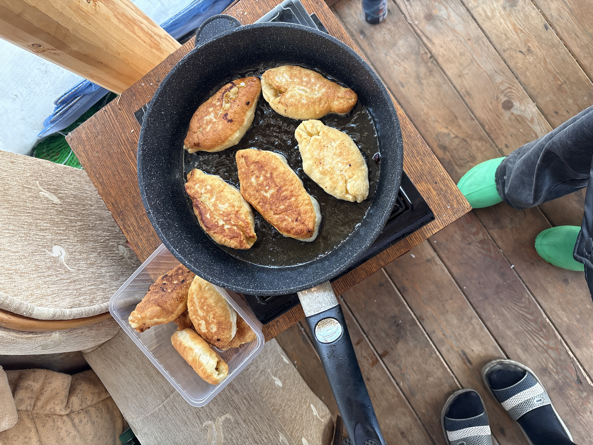 This overhead image depicts an outdoor cooking scene, likely on a wooden deck or porch, with an emphasis on food preparation. At the center, a black frying pan filled with cooking oil holds six golden-brown patties or pies, sizzling on a portable gas stove. To the immediate left, a clear plastic container stores four additional cooked patties, indicating a batch being prepared or served.
The cooking setup rests on a dark wooden table or surface. To the left of this area, a light wooden beam or post is visible, along with stacked fabric items, possibly cushions or blankets, and a glimpse of blue mesh material, perhaps a net.
The bottom right of the frame shows the lower legs and feet of two people. One person wears dark grey pants and bright neon green slip-on shoes or boots. Another pair of feet, clad in black and white striped sandals with a faint "NIKE" logo, are visible on the rustic wooden floorboards.
The lighting suggests it is daytime. The handle of the frying pan features a silver button with the word "PRESS" inscribed, possibly for a detachable mechanism. The scene portrays a casual, informal cooking activity, possibly enjoyed during leisure time in Zheleznodorozhny, Russia.