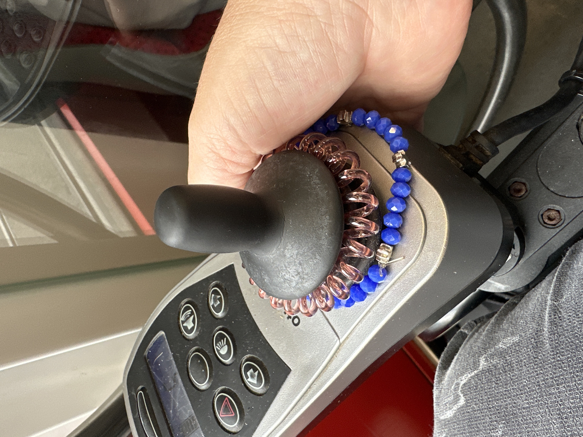 A hand is shown resting on the joystick of a powered wheelchair. The joystick is black and has a coiled hair tie wrapped around its base. A blue beaded bracelet is also wrapped around the base of the joystick. The wheelchair's control panel is visible in the background, with several buttons and a small digital display. The scene appears to be indoors, possibly in a home or medical facility, but there is no specific indication of the city of Krefeld, Germany.