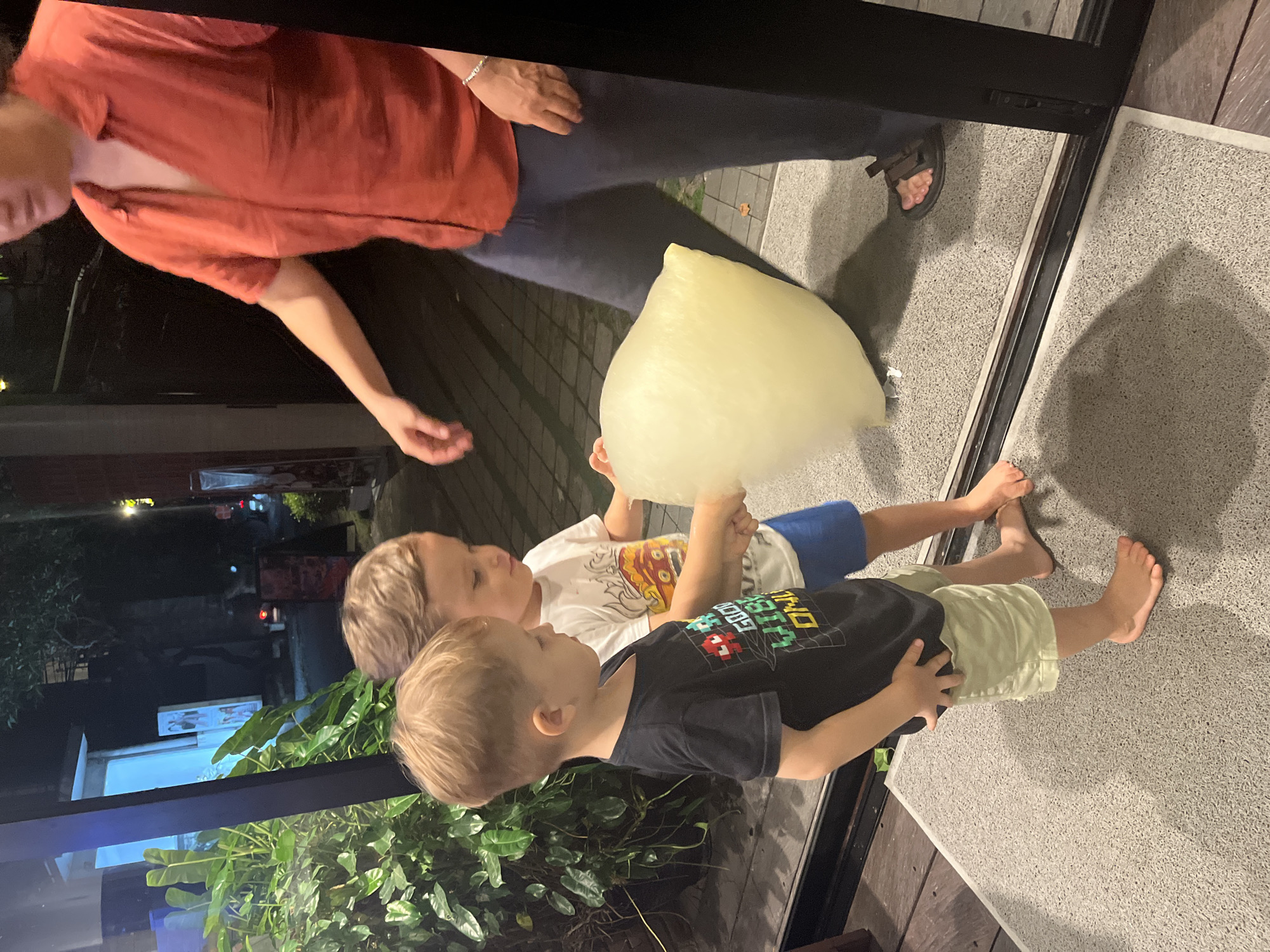 Two young boys, appearing to be brothers, are standing on a grey mat in front of a glass door. The boy on the left is wearing a black t-shirt with a pixelated graphic and light green shorts, while the boy on the right wears a white t-shirt with a colorful graphic and blue shorts. They are both holding a large, amorphous, pale yellow mass, possibly cotton candy. A person in a reddish-orange shirt and dark pants stands behind them, their hand outstretched towards the cotton candy. The background shows lush green foliage, a building with blue lighting, and what appears to be a street scene at night, suggesting an urban or semi-urban environment in North Kuta, Indonesia. The lighting is dim, indicating it is likely evening or night.