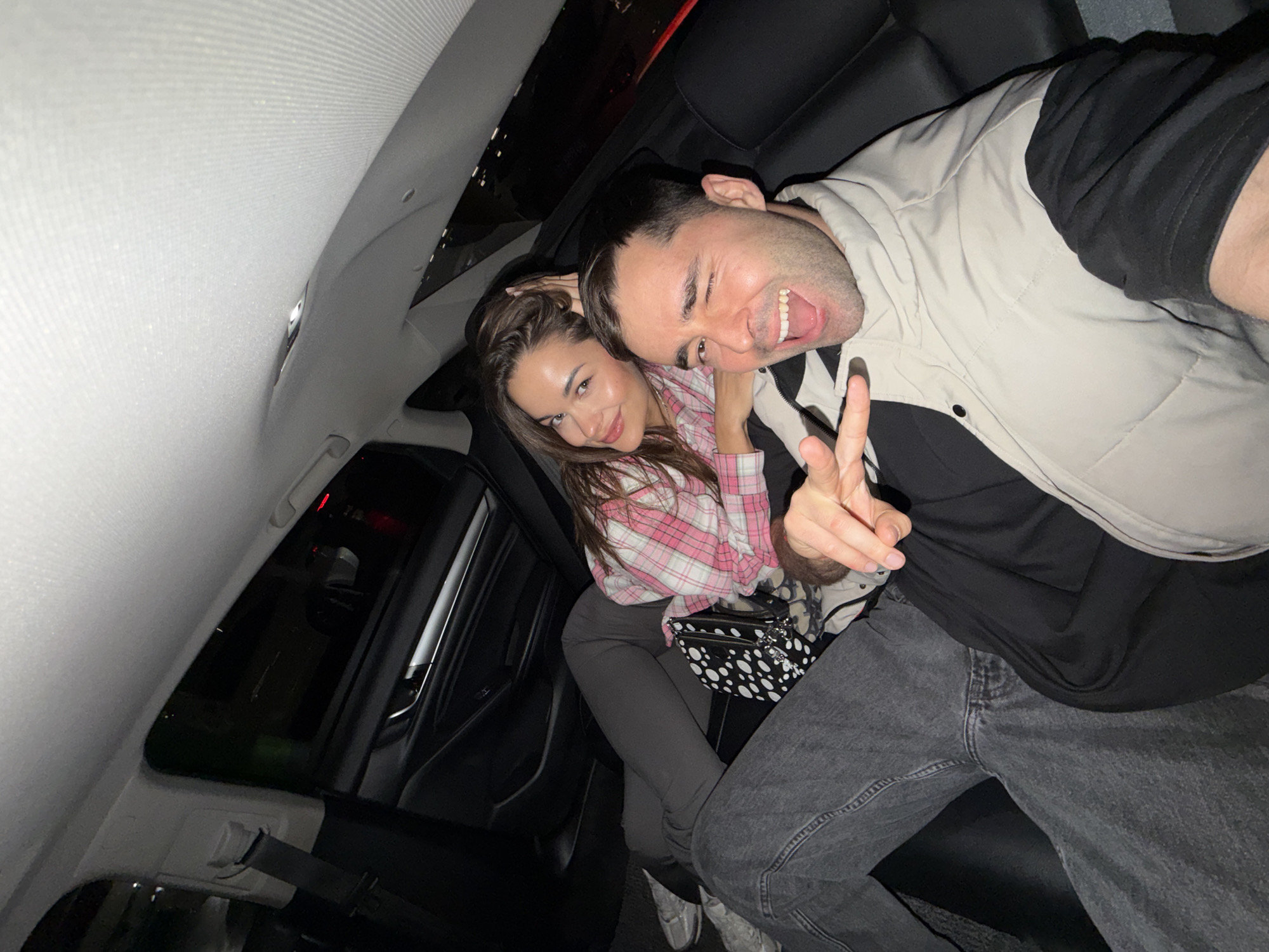 A photograph shows a man and a woman in a car, presumably taken from the backseat. The man is facing the camera and holding up a peace sign with his right hand, with an open-mouthed, energetic expression. He is wearing a black shirt, a light-colored puffy vest, and gray jeans. The woman sits beside him, looking at the camera with a slight smile. She is wearing a pink plaid shirt, gray pants, and white shoes. She has a small black and white purse. The interior of the car is visible, with a sunroof and black leather seats. Based on the visual information, there is no indication of the location being Langfang, China.