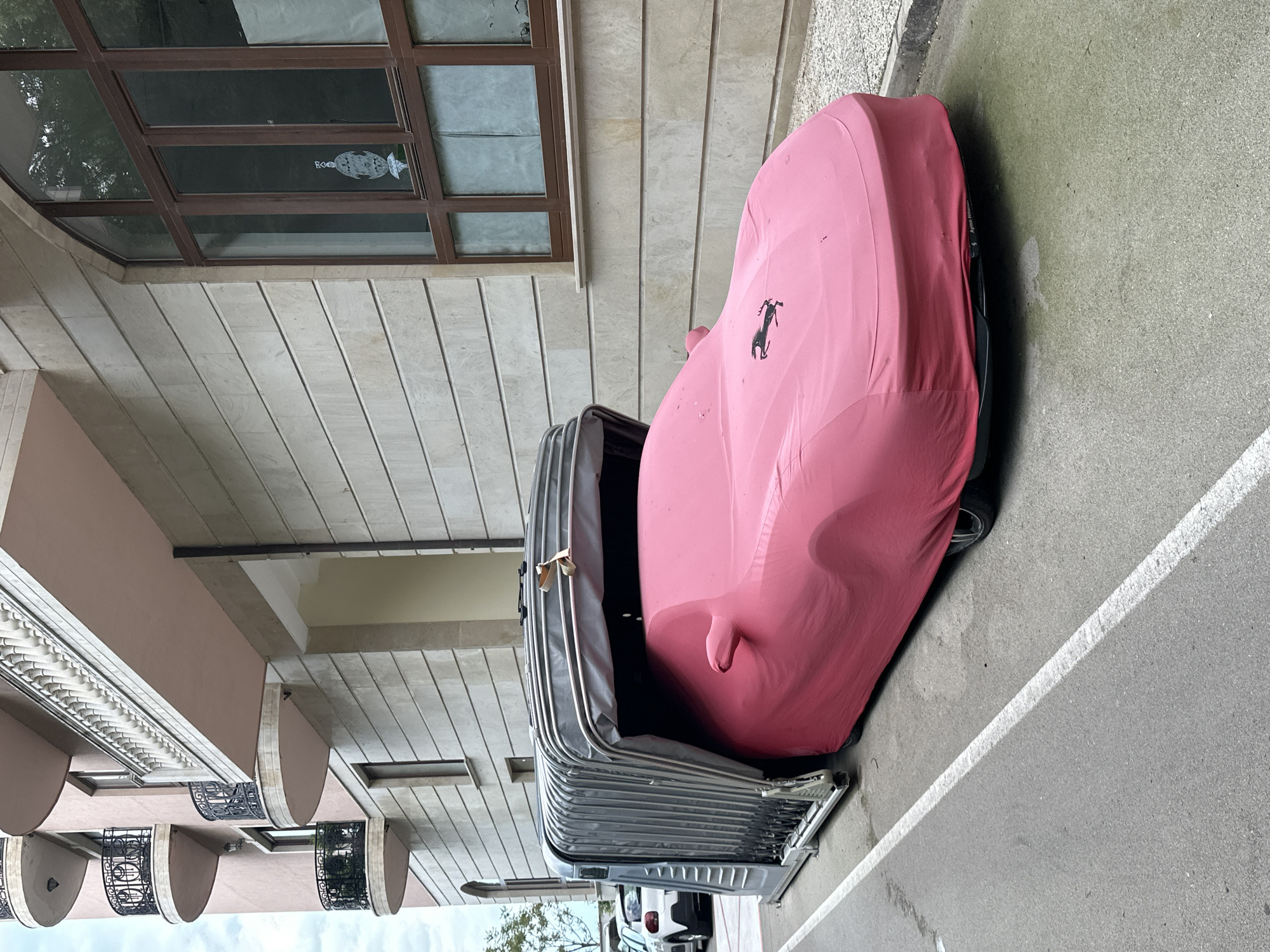 The image shows a pink-covered car parked on a street in Varna, Bulgaria. The car is parked next to a building with stone siding and a window with a decorative window decal. A vehicle storage pod is to the left of the car. The road has a white line marking.