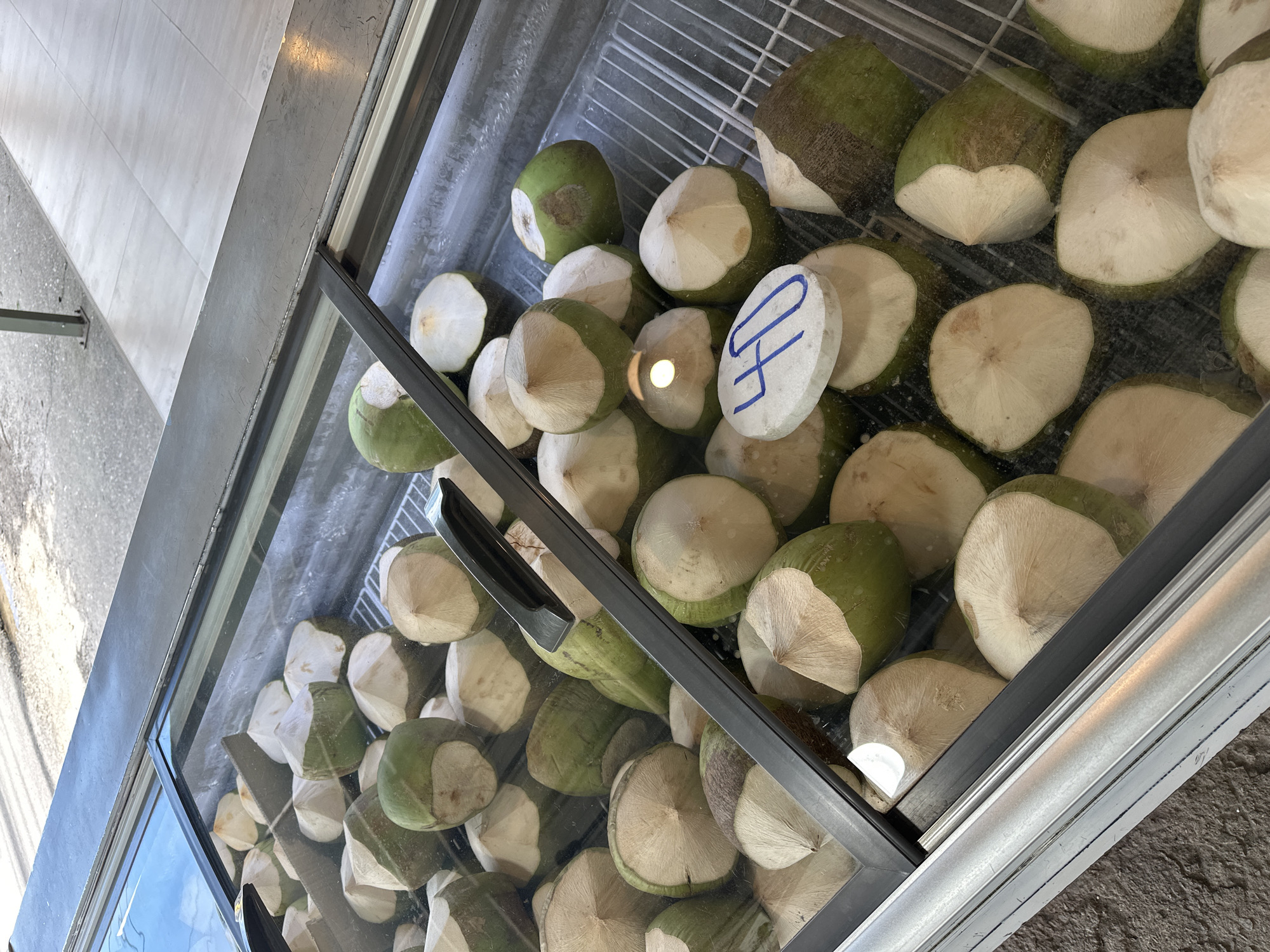 A refrigerated display case is filled with green coconuts that have been partially peeled, revealing the white flesh underneath. The coconuts are arranged on a white grate. A black metal handle or divider is positioned diagonally across the display case. A round white object with the number "40" written in blue marker is placed on top of some of the coconuts in the foreground. The display case is made of glass and metal. In the background, a tiled sidewalk and a textured ground surface are visible. The lighting suggests an outdoor or semi-outdoor environment.