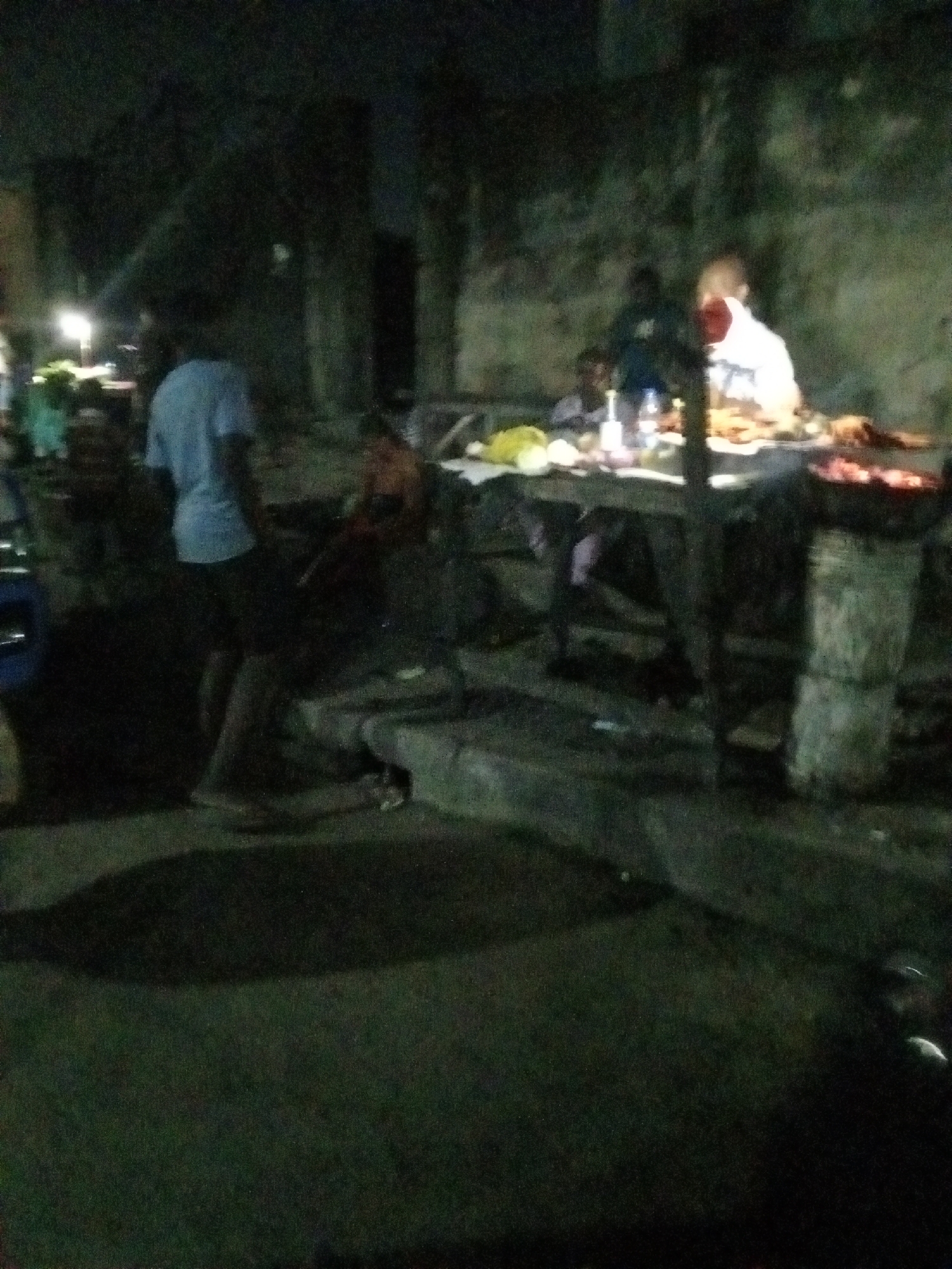 At night, the scene depicts an outdoor food vending stall in Ado, Nigeria. A man stands behind a table laden with cooked food and several bottles of drink. He appears to be serving or preparing orders. A charcoal grill stands nearby, with glowing embers visible. Another man is standing next to a parked car, who is wearing a blue shirt. Additional people are seated and standing around the stall. The surroundings are dimly lit, with dark shadows cast on the ground and a building in the background. The environment is urban with a paved area around the food stall.