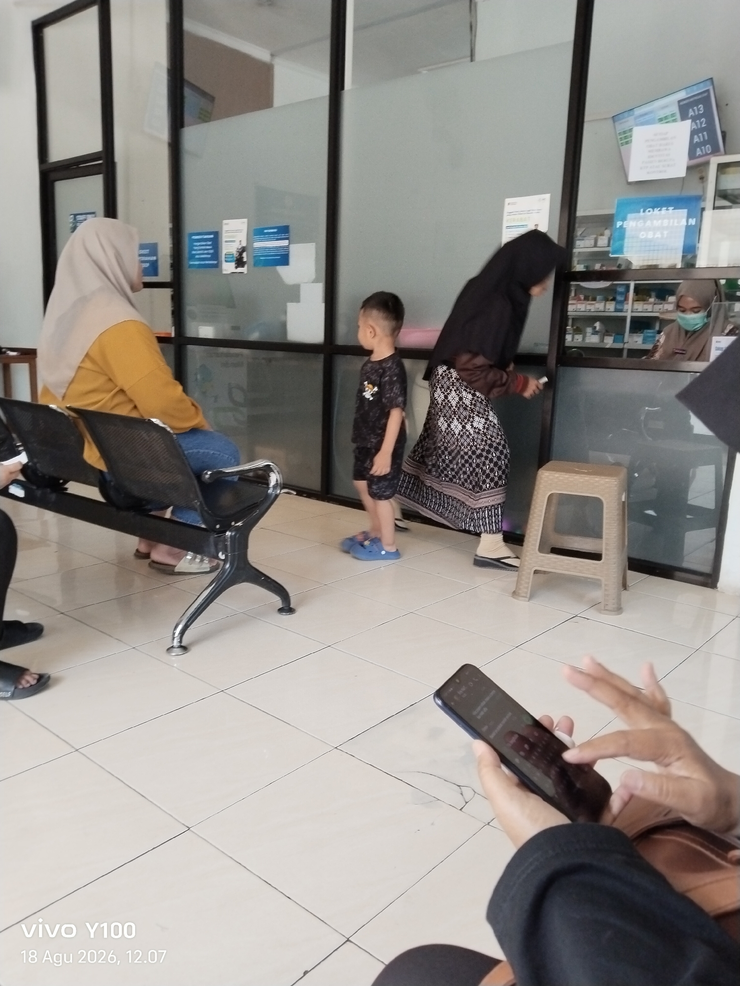 The image depicts a waiting area, likely at a healthcare facility in Patokan, Indonesia. In the foreground, a person's hands are visible, holding a smartphone and interacting with its screen. The background reveals a seating area with several individuals. A woman wearing a light-colored hijab and a yellow shirt is seated on a dark bench. Next to her, a young child in a dark patterned shirt and shorts stands. Further back, another woman in a dark hijab is interacting with a pharmacy counter. Behind the counter, a person wearing a face mask and a brown uniform is visible, along with shelves stocked with medications. A digital display above the counter shows information with text that includes "A13", "A12", "A11", and "A10". Another sign above the counter reads "LOKET PENGAMBILAN OBAT". The environment appears to be indoors, with tiled flooring and glass partitions. The lighting is typical of an indoor setting, with some reflections visible on the glass surfaces. A timestamp in the bottom left corner indicates "vivo Y100" and "18 Agu 2026, 12.07".