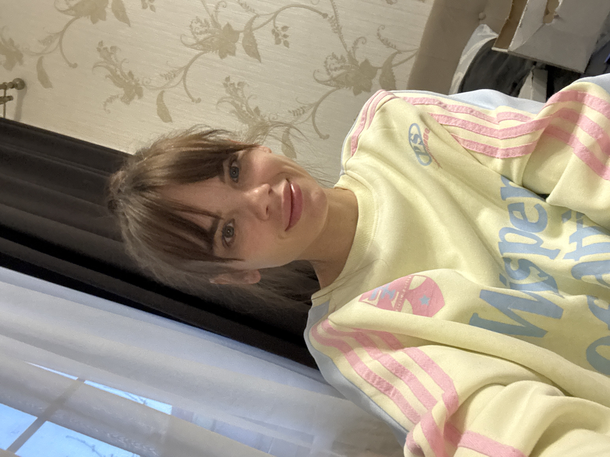 A portrait-style selfie captures a young woman indoors in Rostov-on-Don, Russia. The image is rotated, with her head tilted to the left. She has light brown hair with bangs, blue eyes, and a gentle smile, looking directly at the camera. She is wearing a pale yellow sweatshirt featuring pink stripes on the sleeves and light blue lettering on the front. Visible text on the sweatshirt includes "PS" (or a similar logo) on the left shoulder and parts of a larger word, possibly "Wisper," in a light blue script across the chest.
The background shows a light-colored wall adorned with a delicate floral or damask wallpaper pattern in a slightly darker beige tone. To her left, a window with horizontal white blinds is partially open, revealing a clear blue sky and bare tree branches, suggesting a daytime setting in late autumn, winter, or early spring. A dark brown element, possibly a window frame or furniture, borders the window. Hints of light-colored furniture or bedding are visible in the upper right background. The overall mood conveyed by her expression is calm and pleasant.