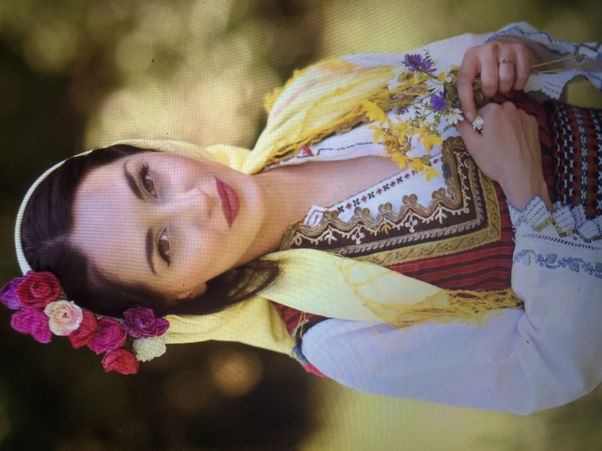 The image depicts a young adult female, likely in her 20s or 30s, positioned centrally and looking directly at the camera with a slight smile. She has dark hair, dark eyebrows, and brown eyes with visible eyeliner and dark mascara. Her lips are painted a matte red. She is wearing traditional attire, commonly associated with Bulgarian folk costumes. Her head is adorned with a pale yellow headscarf draped over her dark hair, with a floral circlet or wreath of artificial flowers (predominantly shades of fuchsia, magenta, red, and cream/white) positioned above her right ear. She wears a white long-sleeved blouse with intricate blue and green embroidery patterns on the cuffs. Over the blouse, she has a dark-colored vest featuring ornate gold and brown embroidery around the neckline and chest, covering a striped red and dark fabric garment. A woven belt or apron with red, black, and white geometric patterns is visible around her waist. Her hands, positioned at the lower center of the frame, gently hold a small, informal bouquet of mixed wildflowers, including yellow, white, and purple blooms. A simple silver-toned ring with a small, clear stone is visible on the ring finger of her left hand. The scene is set outdoors against a softly blurred background of green and yellow foliage, indicating natural light. While the attire strongly suggests traditional Bulgarian culture, the specific geographic location in the city of Shumen, Bulgaria, cannot be visually confirmed from the image provided.