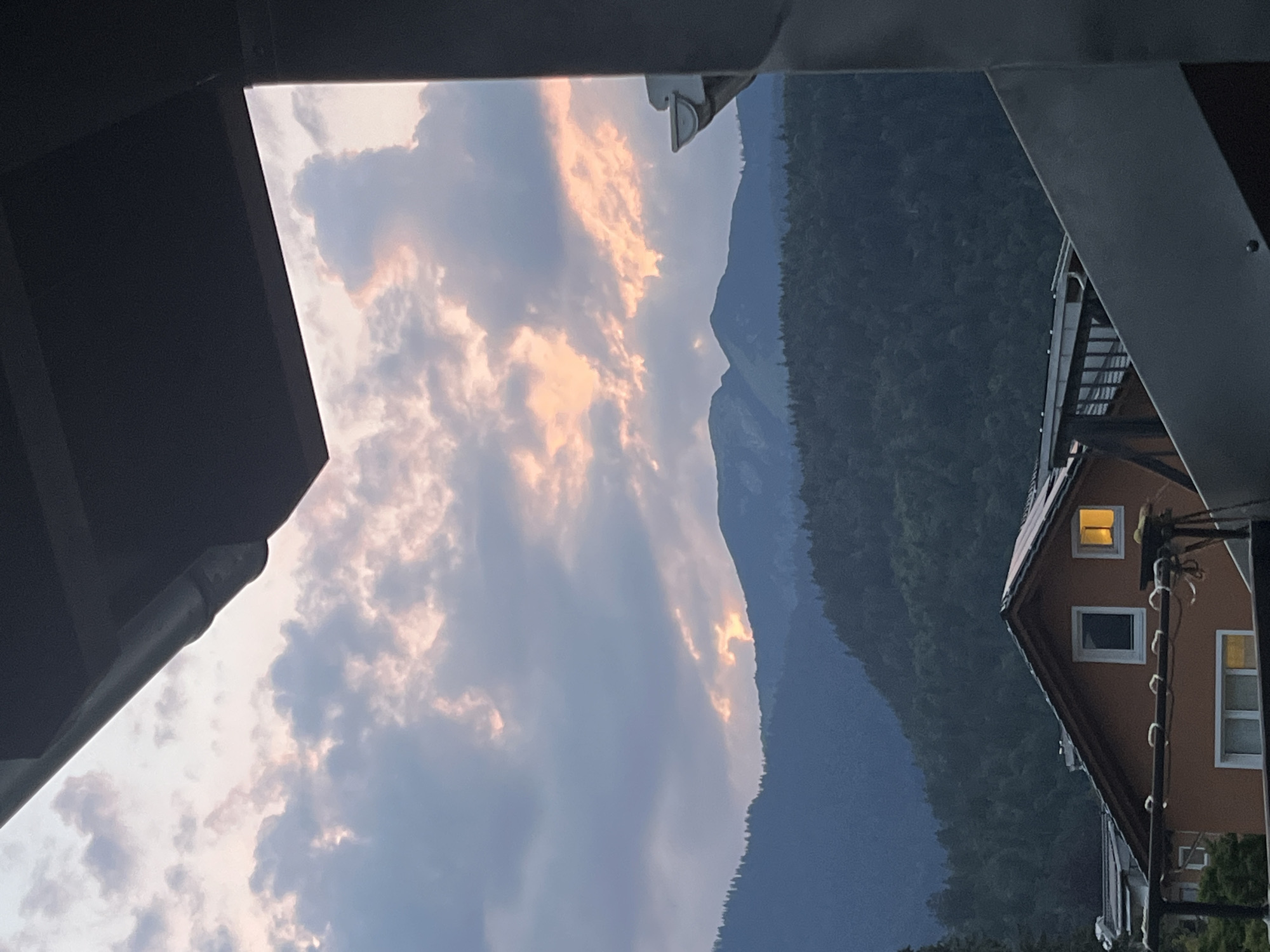 The image shows a view of mountains and a building at dusk. The sky is filled with clouds, some of which are illuminated by the setting sun, casting a warm, orange glow. The mountains in the background are covered in dense green forest, with their silhouettes fading into the twilight. In the foreground, a portion of a building is visible, with windows that are lit from within, suggesting it is a residential or hotel building. The architecture appears to be somewhat rustic, with an orange-brown facade and visible structural elements. The scene is framed by dark, angular structures in the foreground, possibly an overhang or a balcony, which partially obscure the view. The time of day is clearly evening, as indicated by the soft lighting and the illuminated windows. The location is identified as Garmisch-Partenkirchen, Germany, a town known for its alpine scenery. The overall mood is peaceful and serene, capturing the beauty of the natural landscape at the end of the day. There is no visible text in the image.