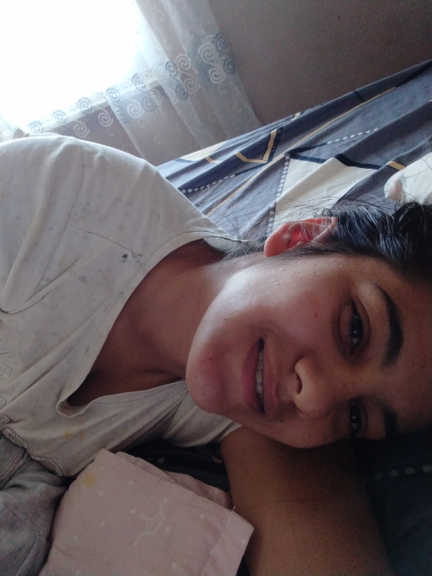 A person lies on a bed with a patterned duvet cover, facing the camera and smiling. They are wearing a light-colored shirt. A window with sheer, patterned curtains is visible in the background. The location is unknown.