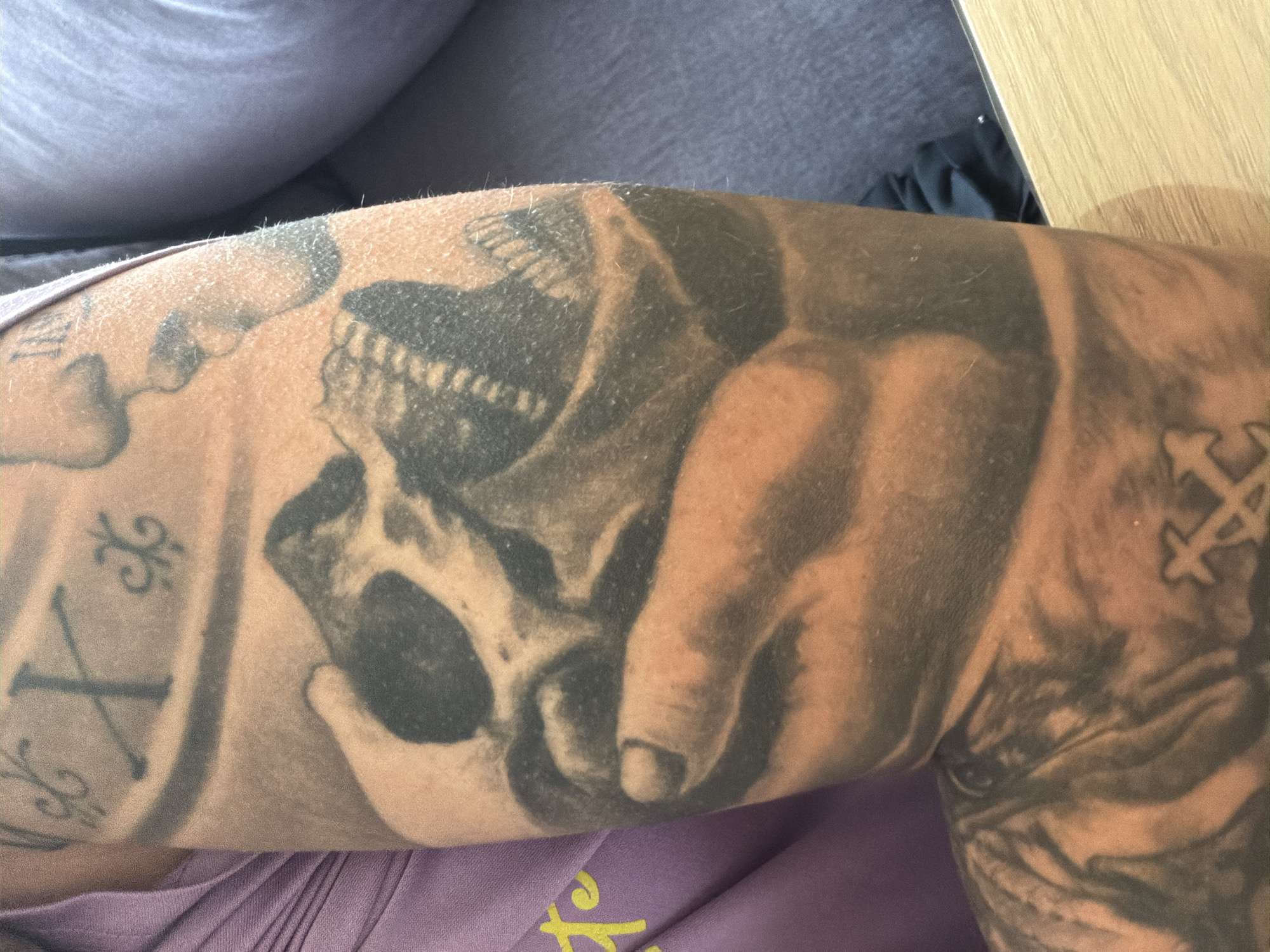 A close-up shot captures a heavily tattooed arm, likely in an indoor setting in Shumen, Bulgaria. The central and most prominent tattoo depicts a detailed human skull, with visible eye sockets, nasal cavity, and teeth, being held from below by a human hand.
To the left of the skull and hand design, a tattoo features a clear Roman numeral 'X' (ten) positioned within a vertical band, flanked by ornate, scroll-like decorative patterns above and below. Further left, faint and darker tattooed areas are visible, blending into the skin.
On the right side of the arm, another tattoo showcases a symbol that appears to be two crossed 'X' shapes or a similar geometric pattern, transitioning into what seems to be a tiger or animal stripe design further to the right.
The arm rests on a piece of purple fabric, possibly clothing or a blanket, which has a blurry yellow design at the bottom. In the upper right background, a light wooden surface, likely a tabletop, is visible next to a dark grey fabric. The lighting indicates an indoor environment. No specific activity is taking place, and no emotions are discernible from the partial view of the person.