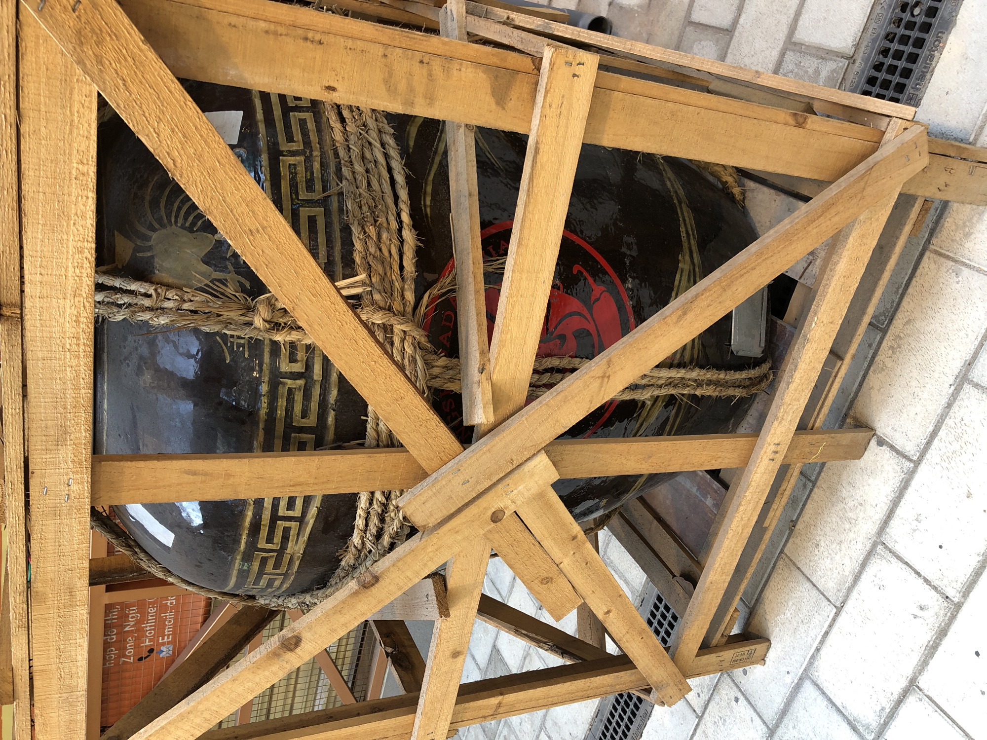 In Đà Nẵng, Vietnam, a large, dark, decorative urn is secured within a wooden crate for transport. The urn features intricate gold Greek key patterns and a stylized rooster motif. Thick ropes bind the urn to the crate, highlighting its careful packing. The crate itself is constructed from rough-hewn wooden planks, creating a protective cage around the valuable item. The background suggests an urban setting, with a tiled pavement and a drain grate visible. A small sign with Vietnamese text, "hộp dẻ, thi. Zone, Ngũ. Hotline:, Email: de," can be seen behind the crate, indicating a local business or service.