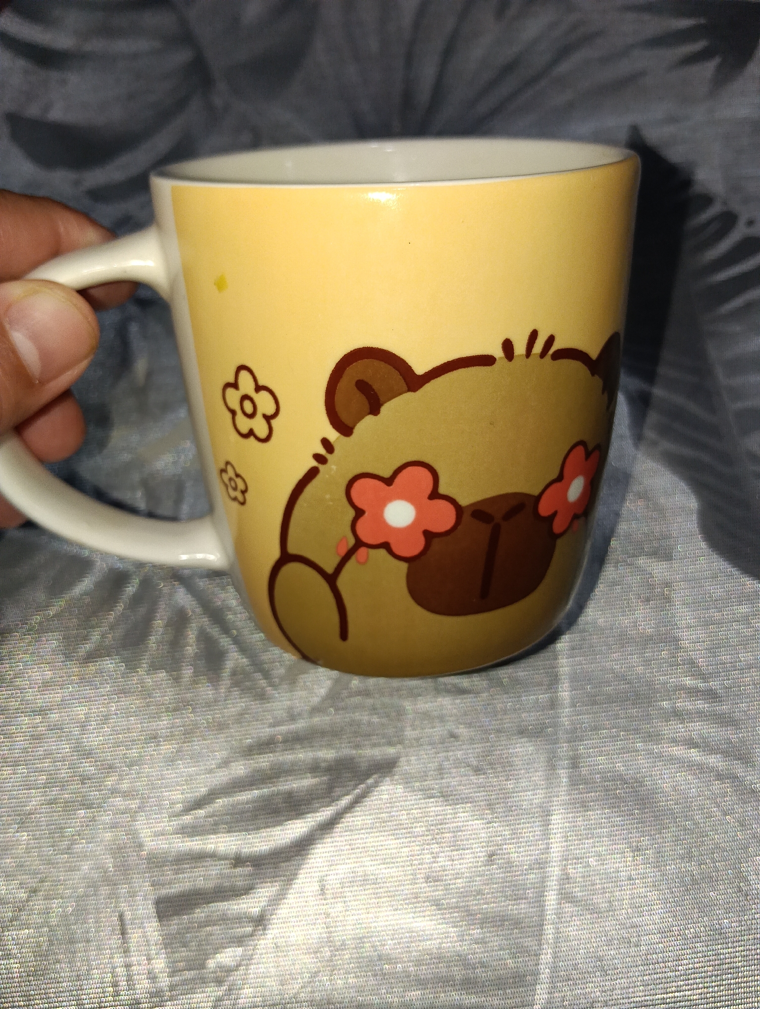 A close-up, eye-level shot shows a yellow mug with a cartoon bear design held by a person's hand. The mug is positioned in the center of the frame, with the bear's face prominently displayed. The bear has large brown eyes, small ears, and a button nose. Pink flowers with white centers are depicted on the bear's cheeks. The mug is made of ceramic and has a white handle. The background consists of a gray, textured surface, possibly a fabric or tablecloth, with some light reflecting off it. The image is well-lit, with a shadow cast to the right of the mug. The image was taken in Kuching, Malaysia.