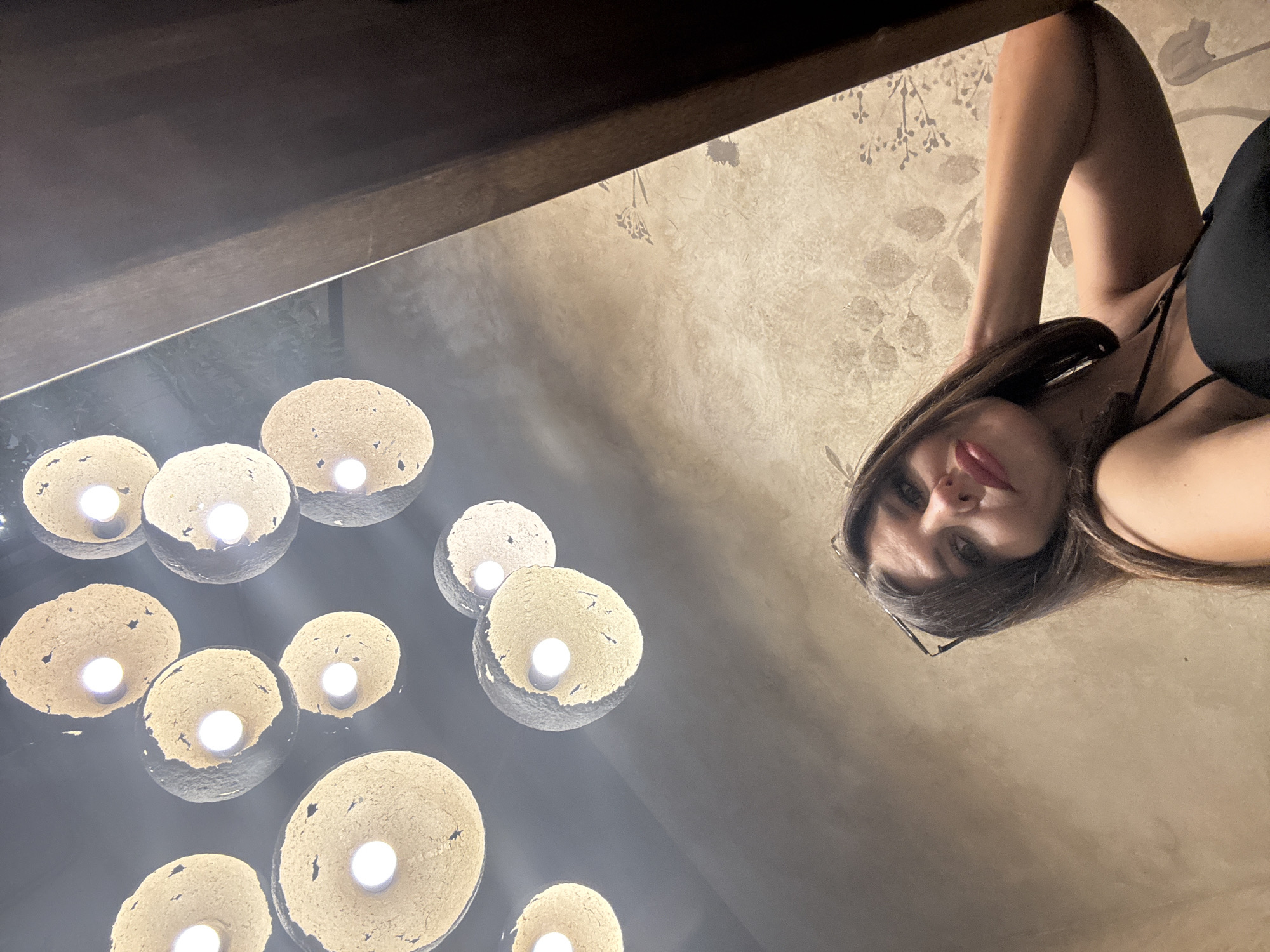 An upward-angled, indoor selfie shows a woman with dark hair and glasses pushed up on her head, wearing a black top. She is posing with her hand on her neck, looking at the camera. The background features a textured, patterned wall and a cluster of decorative pendant lights with exposed bulbs. The scene is dimly lit, with the lights providing the primary illumination.