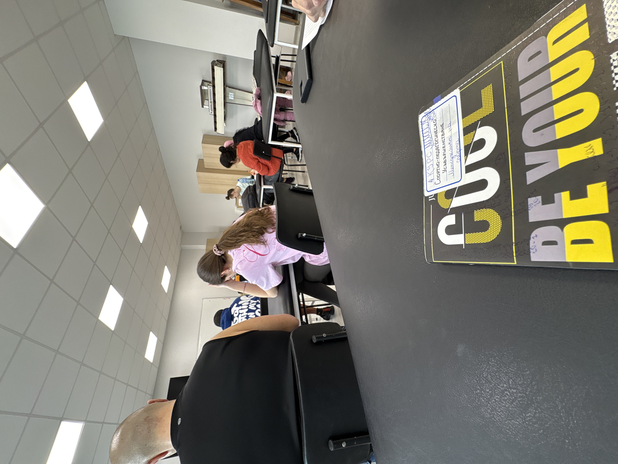 The image shows a classroom in Sofia, Bulgaria. Students are seated at desks, presumably during a lecture or study session. A bald person in a black shirt is visible in the foreground. On a desk is a notebook with the text "BE YOUR... GO UP" and a sticker with Cyrillic text indicating "4th course, sports-pedagogical... specialized in Sports". The room is well-lit with fluorescent ceiling lights and has a neutral color scheme. Other students are seen seated, some wearing casual clothing. The classroom appears to be in a school or university setting.