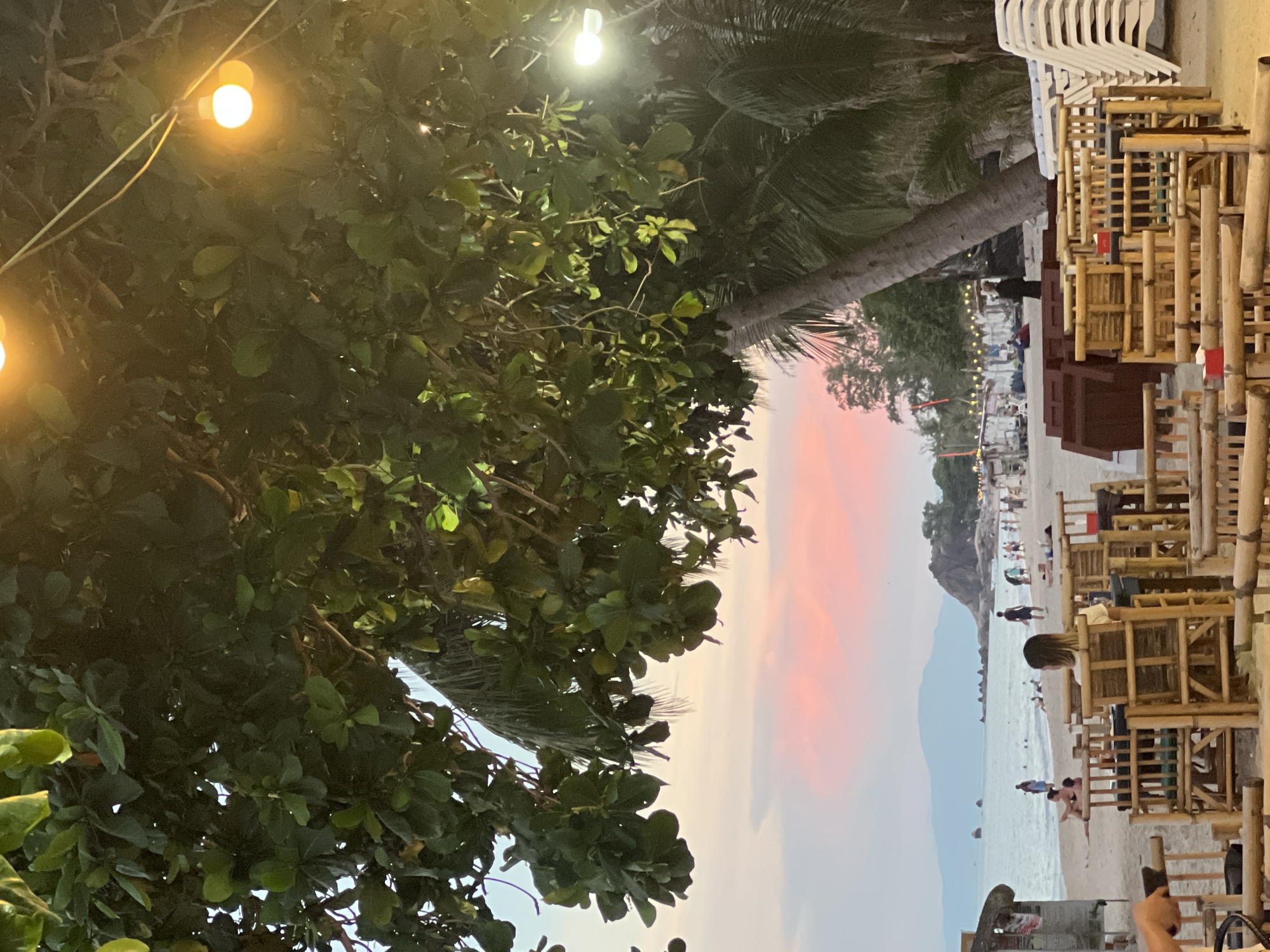 The image depicts a beachfront scene at Baan Lamai, Thailand, likely during the late afternoon or early evening. The foreground is dominated by bamboo seating arrangements and tables, suggesting an outdoor dining or seating area. A woman with dark hair, wearing a light-colored top, is seated facing away from the camera at one of these tables. Beyond the seating area, a sandy beach stretches towards the ocean. Several people are visible on the beach, some walking, others relaxing. The ocean is calm, and in the distance, mountains and rocky outcrops can be seen. The sky exhibits a soft pink and orange hue, indicative of a sunset. Lush green foliage from trees overhangs the scene, and string lights are visible in the background, casting a warm glow. Palm trees are also present, characteristic of a tropical coastal environment. The overall atmosphere appears relaxed and serene.