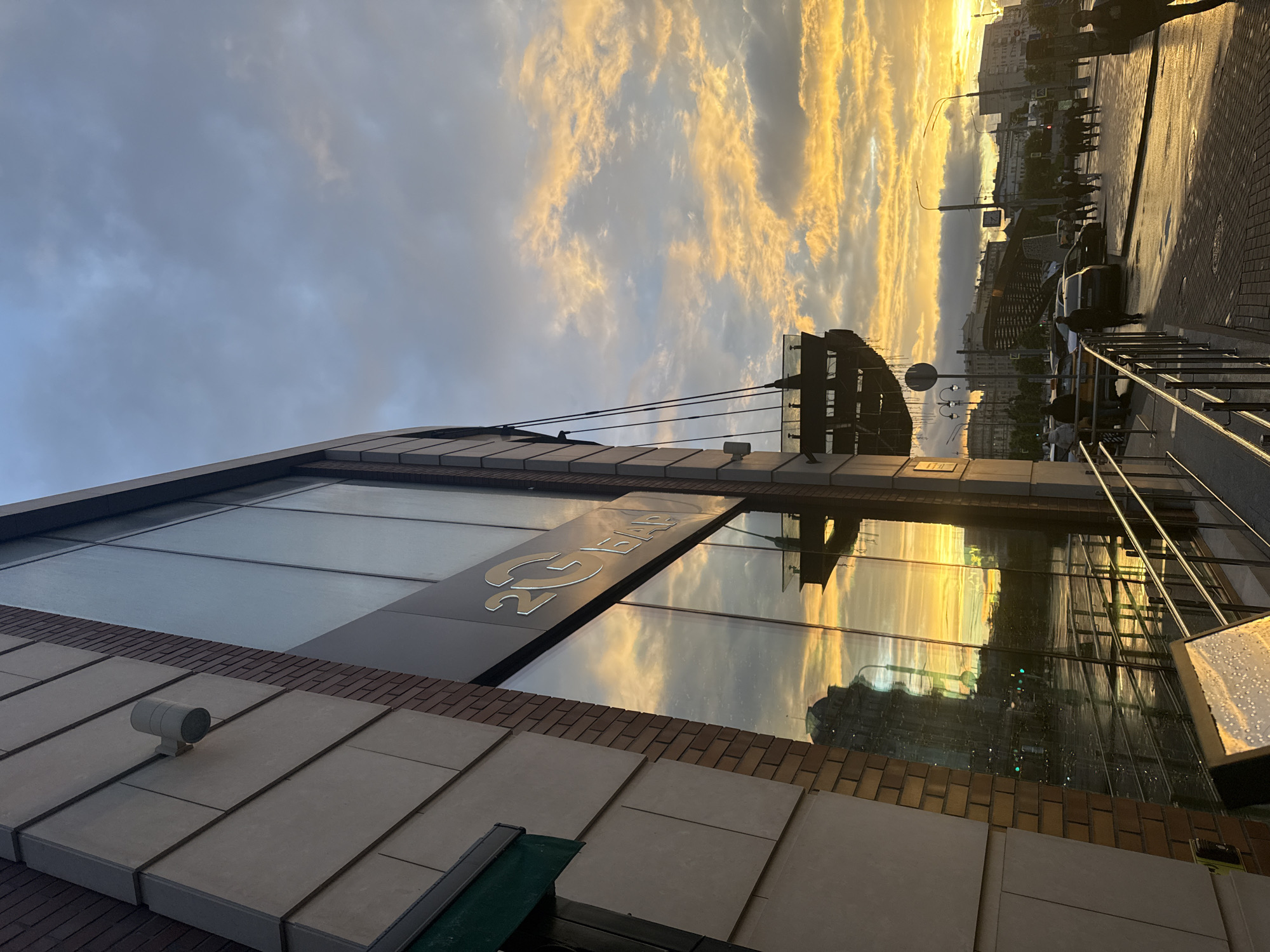 The image depicts a scene in Moscow, Russia, at sunset. The sky is filled with clouds illuminated by the setting sun, creating a golden glow. The reflection of the sky is visible in the glass facade of a building with the sign "2G Bar." The building has a modern design with a combination of brick and glass elements. A security camera is mounted on the building's exterior. In the distance, figures of people can be seen walking along a sidewalk. Cars are parked on the street. A metal structure resembling a bridge or overpass is visible further in the background.