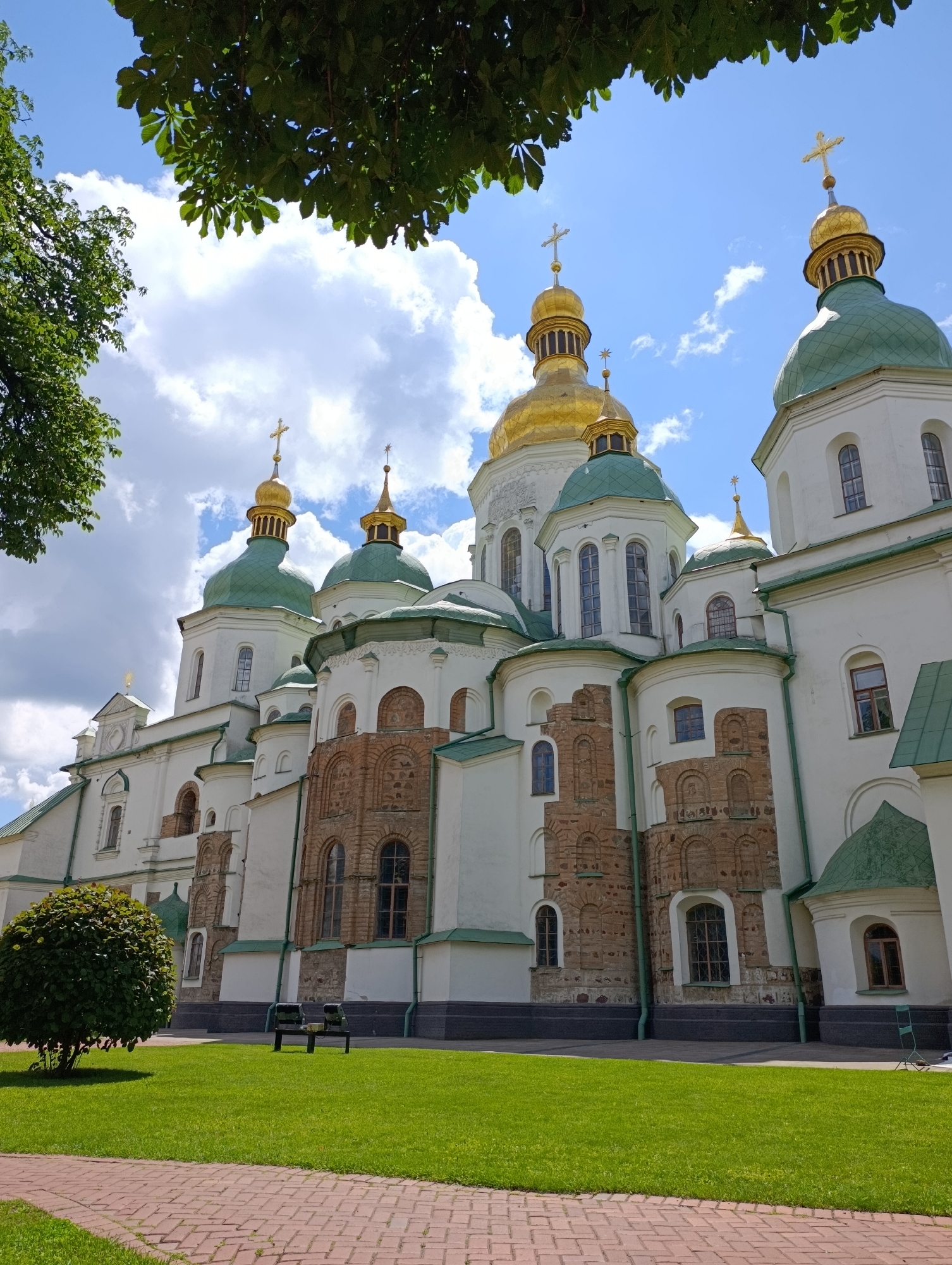 The image depicts the Saint Sophia Cathedral in Kyiv, Ukraine, on a bright, sunny day. The cathedral's white walls are adorned with green domes and gold-plated crosses, characteristic of Ukrainian Baroque architecture. The building complex features a series of onion-shaped domes, some gilded and others green, topped with crosses, reaching towards a bright blue sky dotted with white, fluffy clouds. The lower sections of the cathedral's walls display exposed brickwork with arched windows. In the foreground, a meticulously manicured green lawn stretches out, bordered by a red brick pathway. A small, round, leafy tree stands to the left of the lawn, and a dark-colored bench is visible in the middle, with two seats. The scene is serene and architectural, highlighting the grandeur of the historical landmark.