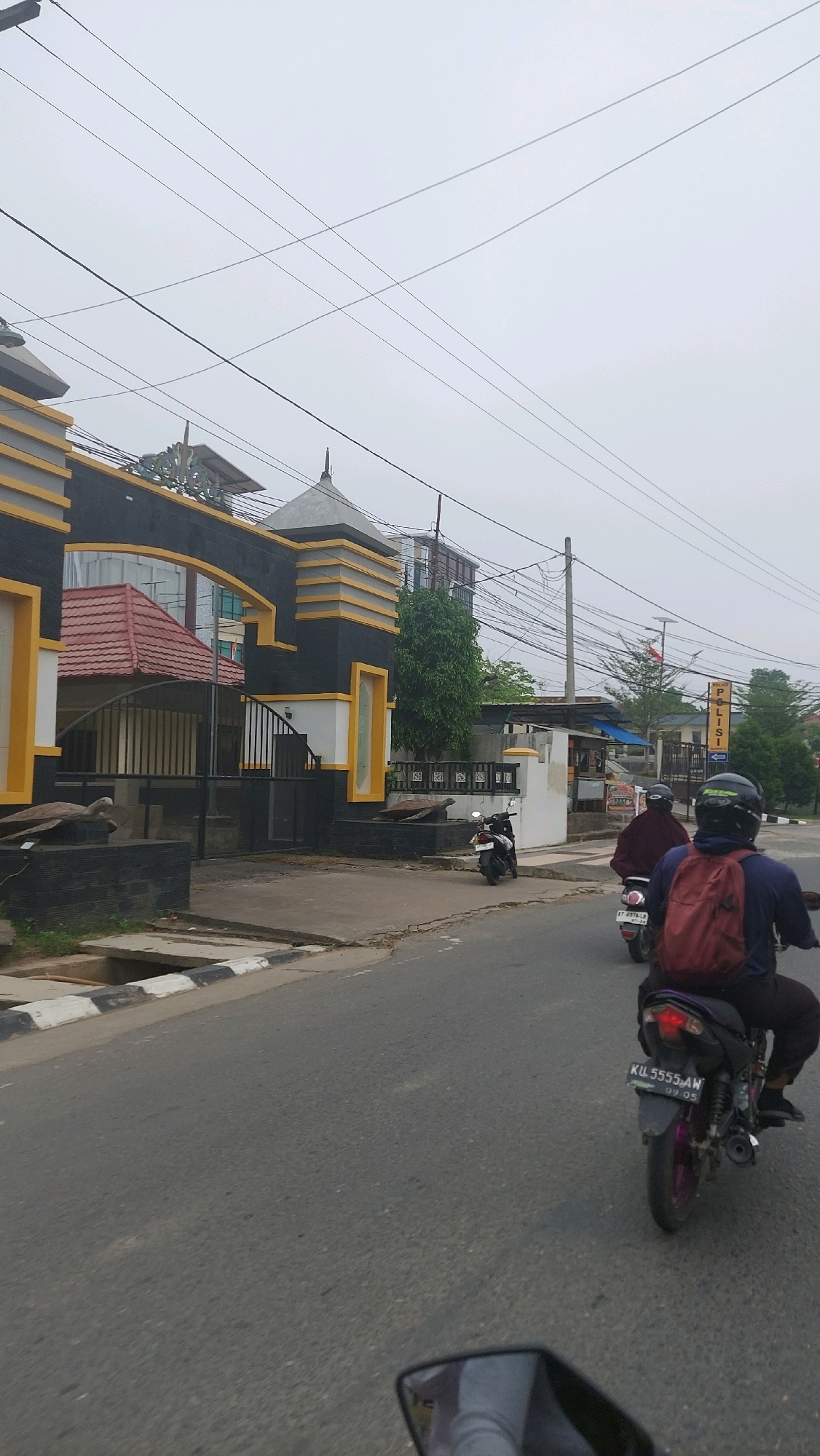 The image depicts a street scene in Sungai Bedungun, Indonesia, with a building on the left and a road in the foreground. The building features a black and yellow architectural style, with a prominent arched entrance. Two turtles are placed as decorative elements in front of the building's entrance. A motorcycle is parked to the right of the entrance.
The road shows signs of wear and tear, with a concrete curb and drainage channel on the left side. The sky is overcast, suggesting cloudy weather.
Several people on motorcycles are present. One motorcycle, viewed from the rear, has a license plate that reads "KUL 5555 AW". The rider is wearing a dark blue shirt and a red backpack. Another motorcycle is ahead of this one, with a rider wearing a dark maroon jacket. A third motorcycle is further ahead and to the left, with a rider whose gender is not discernible from this angle.
In the background, a building with "POLISI" signage is visible, indicating a police station. Overhead, numerous power lines crisscross the sky, a common sight in urban and semi-urban areas. Trees are scattered throughout the scene, and the overall impression is that of a moderately developed area within the city.