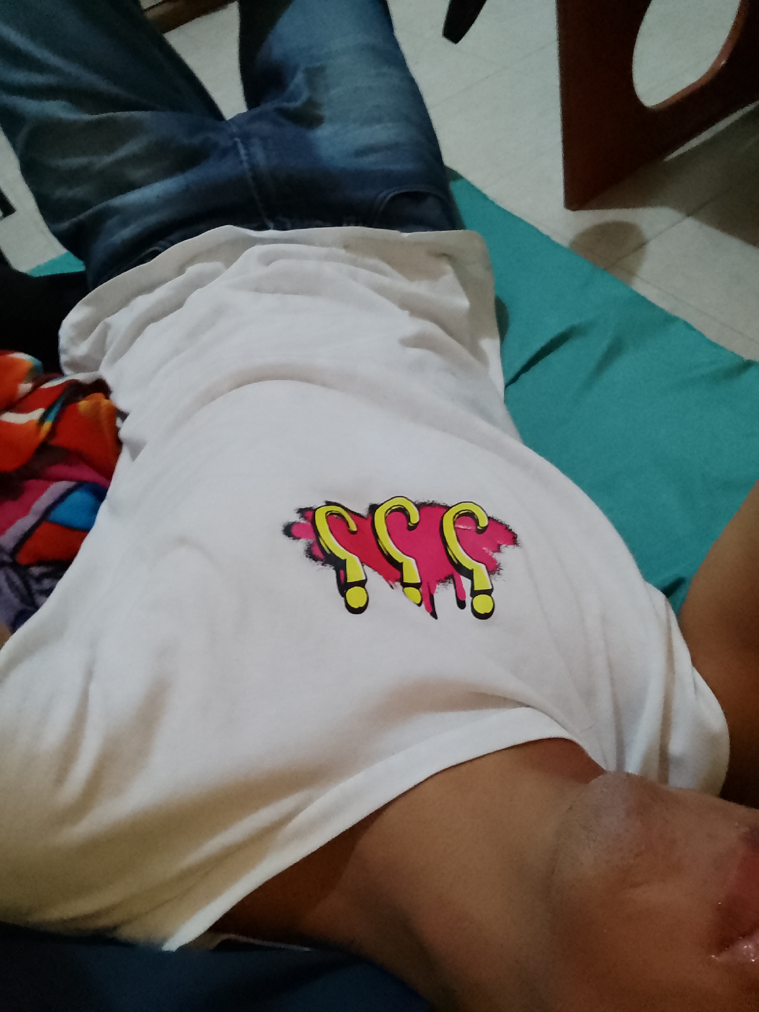 A person wearing a white t-shirt with three dripping pink question mark designs outlined in yellow is lying on a turquoise beanbag. The person is wearing blue jeans. A small wooden table with a circular cutout is in the background. The photograph was taken indoors, likely at eye level, and does not provide identifiable location cues specific to Pampatar, Venezuela. No additional people or actions are apparent.