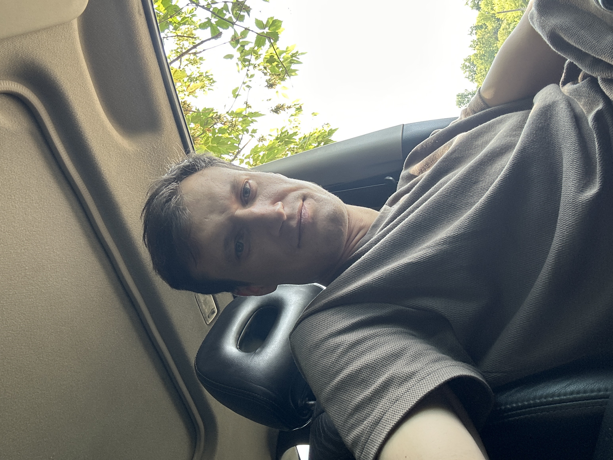 A selfie shows a man sitting in the passenger seat of a car. He is looking at the camera with a neutral expression. The man is wearing a brown t-shirt. The interior of the car is visible, including the headrest of the seat behind him and the sunroof above. Through the car window, trees and foliage can be seen against a bright sky. The image was taken during the daytime. The specific location within Makiv, Ukraine cannot be determined from the visual information provided.