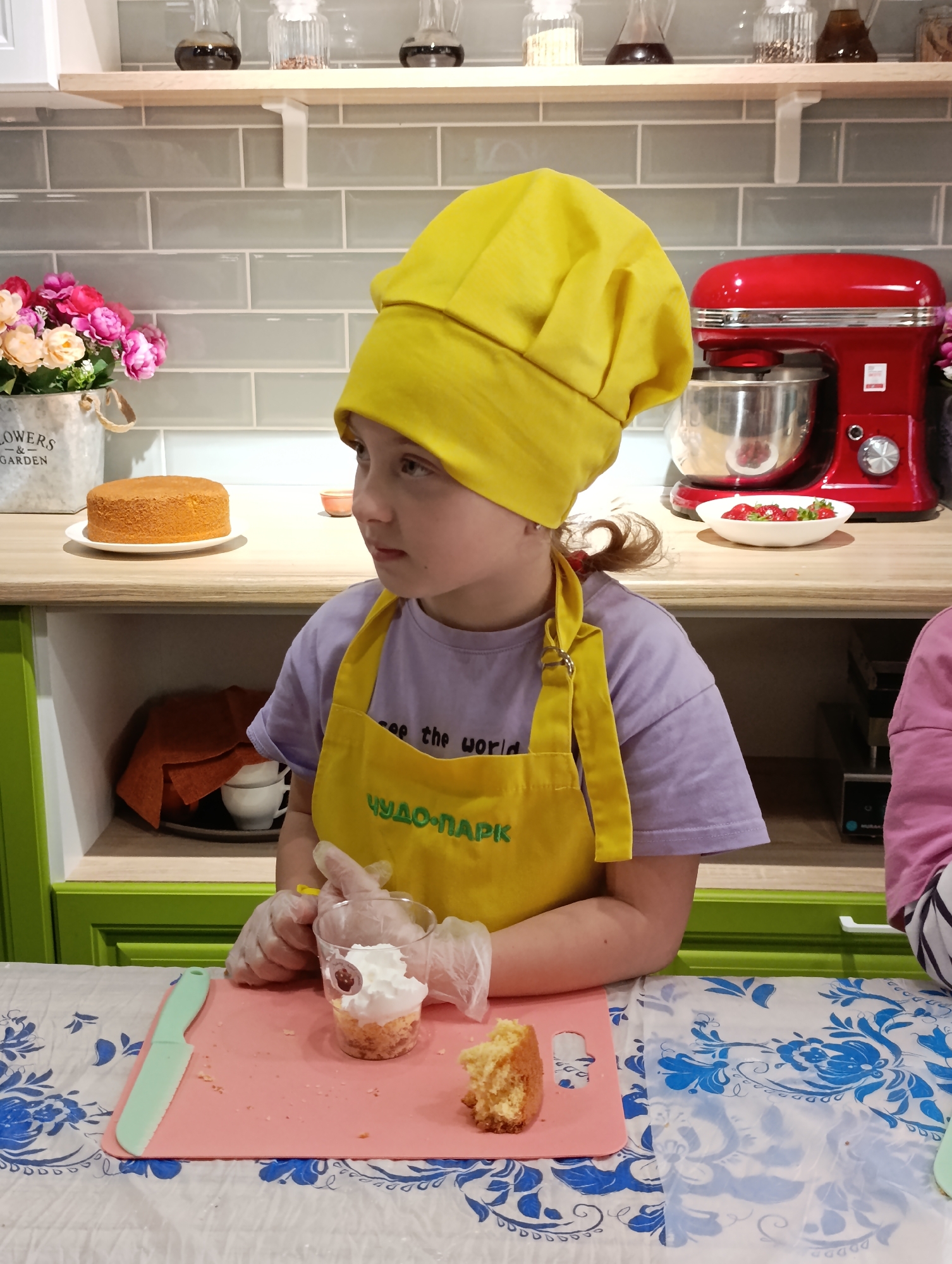 A medium shot captures a young girl in a bright yellow chef's hat and apron, engaged in a baking activity indoors. The girl is wearing a light purple t-shirt under her apron, which features Cyrillic script. She has fair skin, and her gaze is directed slightly to the left. She wears transparent gloves on her hands.
In front of her, on a table covered with a blue and white floral patterned tablecloth, is a small pink cutting board. Atop the board, a clear plastic cup contains layers of cake and whipped cream, and a piece of cake rests beside it. A light green handled knife lies to the left.
Behind the girl, the background includes a kitchen setting with light green cabinets and a countertop covered with a wooden surface and a gray tiled backsplash. A stand mixer, bright red in color, sits on the counter along with a white plate holding strawberries. To the left, there is a pot of pink flowers, and a cake is displayed on a white plate. Jars of varying contents line a shelf above.