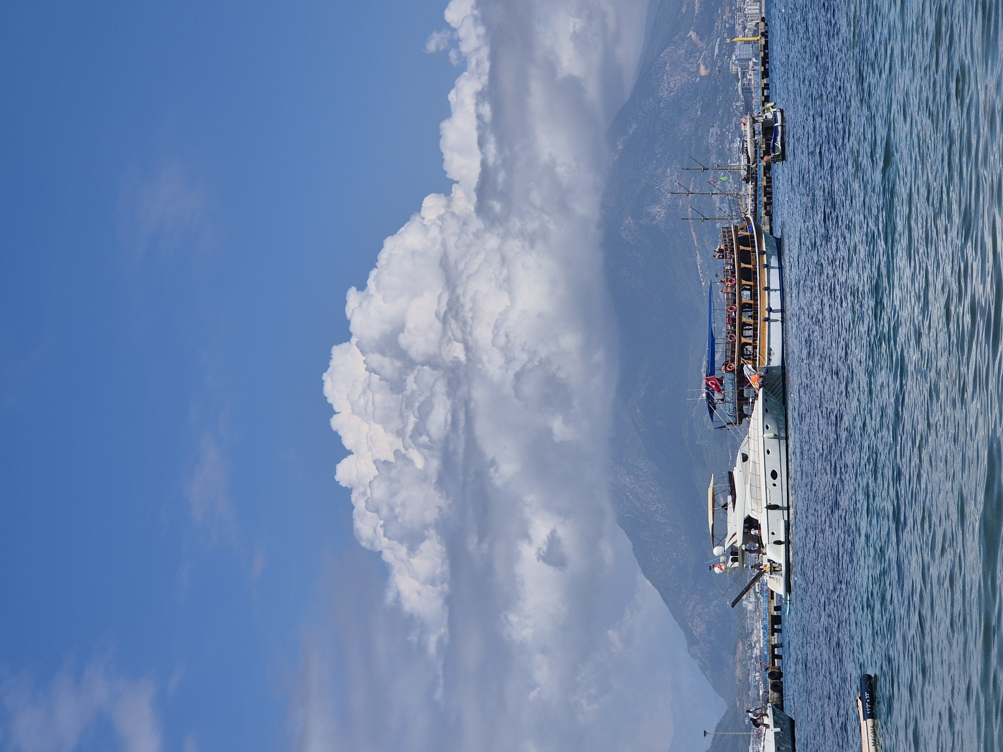 A large, white yacht is docked in the foreground of the image, with several people visible on its deck. Behind the yacht, a larger, traditional wooden tour boat is also docked, crowded with passengers. In the background, a mountain range rises behind the city of Alanya, Turkey, with a large, fluffy cloud formation obscuring the peak of the tallest mountain. The water is a deep blue, with a gentle ripple. A distant pier with several boats and buildings is visible on the right side of the image.