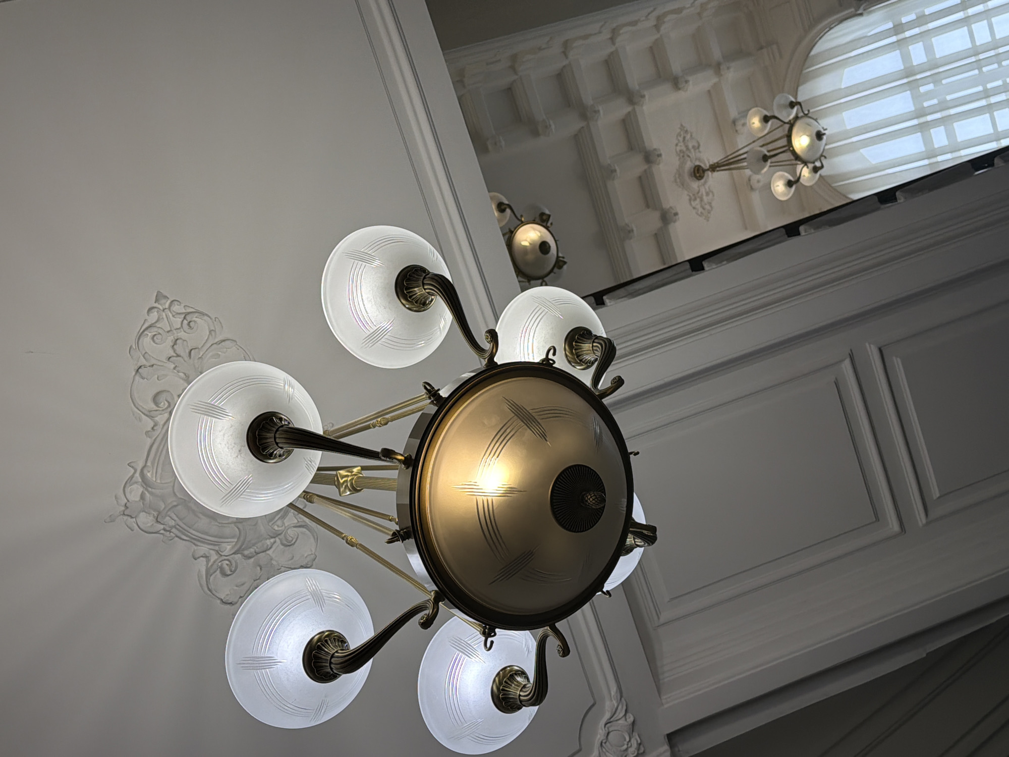 A low-angle shot reveals a light fixture with five frosted glass shades and an opaque central globe, set in a room with decorative molding. The style appears to be vintage. In the background, another light fixture is visible, along with a transom window. The location is likely an interior space within Kazan, Russia, though further information would be needed to confirm.