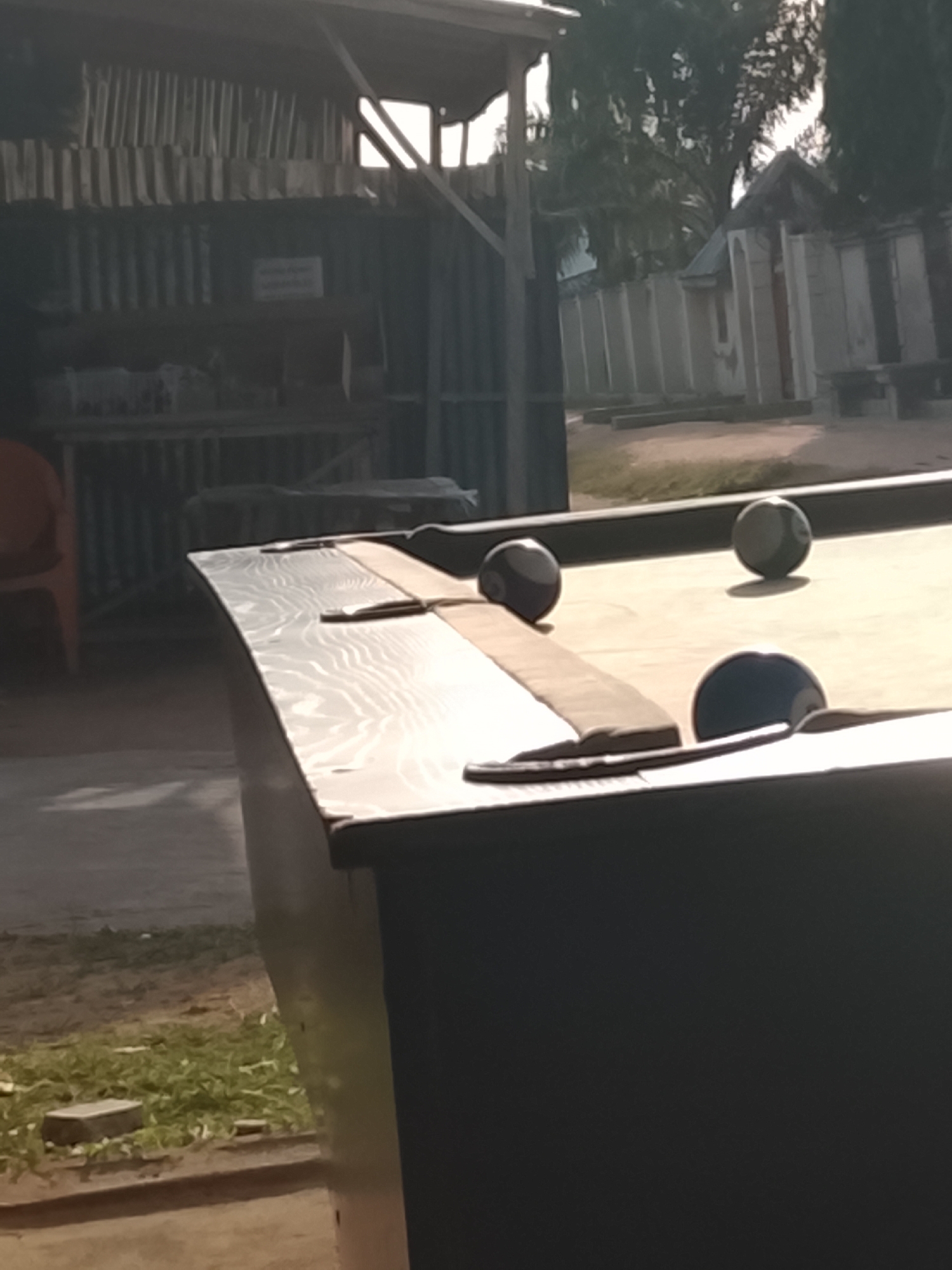 The image shows an outdoor scene, likely in Jalingo, Nigeria, featuring a billiards table with three balls on its surface. The lighting suggests it is daytime, possibly late afternoon given the long shadows. The setting appears to be a casual, perhaps open-air recreational area, with a building or structure in the background that has corrugated metal siding. Trees and other vegetation are visible, indicating an outdoor environment. The billiards table has a dark-colored frame and a light-colored playing surface with a fabric boundary. The balls are dark colored, one of which appears to be partially obscured by the table's edge. The overall impression is one of a quiet moment, possibly before or after a game of billiards. There is no visible text in the image.