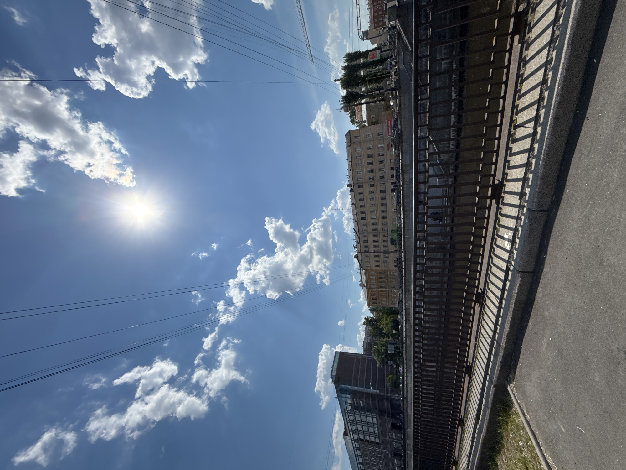 The image captures a bright, sunny daytime scene in an urban environment, likely alongside a canal or river embankment. The sky is a clear blue with scattered white cumulus clouds. The sun is prominently visible in the upper left, shining brightly and casting distinct sunrays. Several thin power lines stretch across the upper part of the sky.
A dark metal railing runs diagonally across the mid-ground from left to right, separating a dark body of water (presumably a canal or river) from a concrete path or sidewalk. The strong sunlight creates long, sharp shadows of the railing's vertical bars onto the path, indicating either morning or late afternoon. To the right of the path is a grey asphalt surface, possibly a road or larger sidewalk.
In the background, a series of multi-story buildings line the water's edge. Their architecture varies; some appear older with numerous windows, while others seem more modern. Green trees are interspersed among the buildings. A construction crane is faintly visible in the upper right background. No people are distinctly visible in the frame, suggesting a calm moment.
Visible text on various buildings includes signs in Cyrillic script, such as "МАГАЗИН" (SHOP), "Ремонт и отделка" (Repair and finishing), and possibly "ПРОДУКТЫ" (PRODUCTS) and "ГАЗЕТЫ" (NEWSPAPERS). A banner or sign with "АРЕНДА" (RENT) is also seen. One sign appears to display "SBERBANK". The bright conditions suggest good weather, and the prominent shadows reinforce a sunny day.