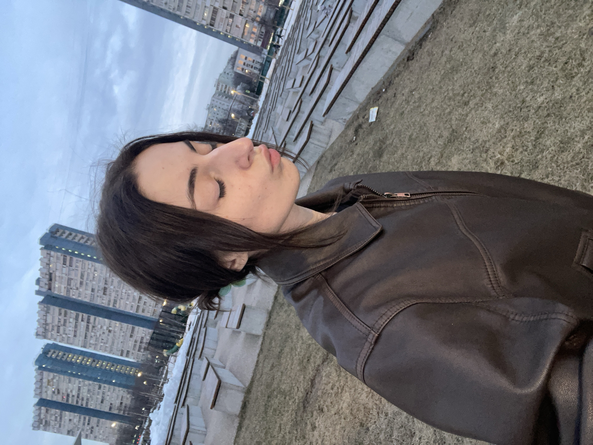 A young female is depicted in a profile view, facing right, with her eyes closed and lips pursed into a kissing gesture. She wears a dark brown leather or faux-leather jacket, and her long, dark brown hair is partially tied back, revealing a light green hair tie behind her left ear. The background features an urban park or public space with extensive tiered concrete seating topped with wooden planks, alongside patches of dry, light brown grass and remnants of snow, suggesting a cold season. In the mid-ground and background, multiple multi-story residential buildings are visible. The most prominent structures are the distinctive "House on Begovaya" complex in Moscow, Russia, characterized by their tall, slab-like appearance, light-colored facades, and prominent blue-paneled sections with numerous windows, some of which are illuminated. The sky is overcast with grey clouds, and artificial lights are visible on buildings and street lamps, indicating low light conditions, possibly dusk or dawn. A small piece of white litter is visible on the ground near the tiered seating.